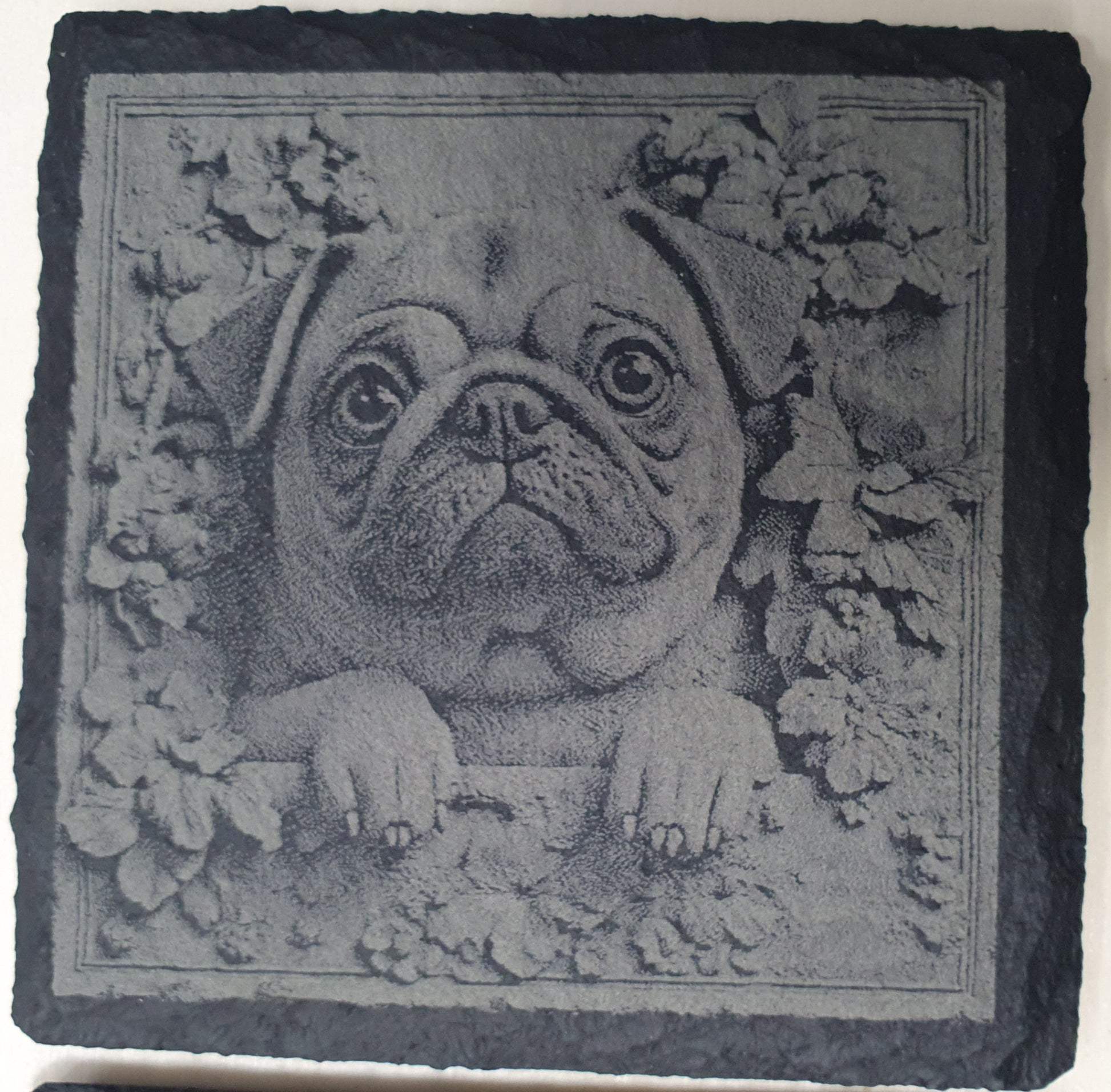 Decorative stone with a pug dog design surrounded by floral elements on a black slate background.