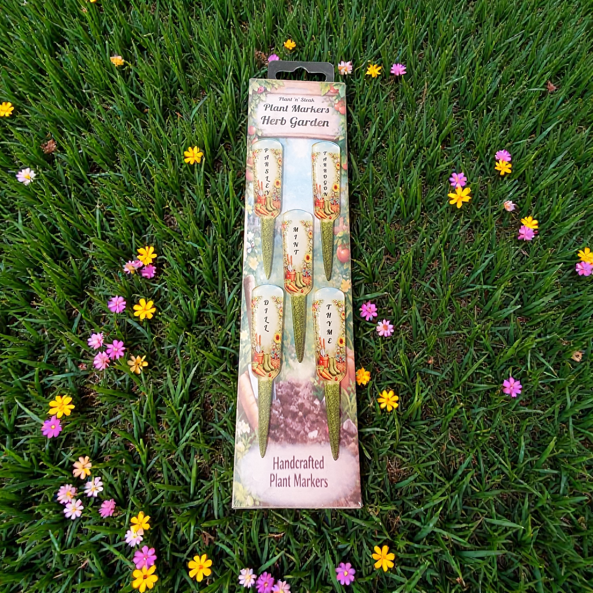Set of 5 handcrafted herb plant markers in retail packaging displayed on green grass with pink and yellow flowers, labelled Parsley, Tarragon, Mint, Dill and Thyme, 10cm acrylic garden stakes made in the UK.