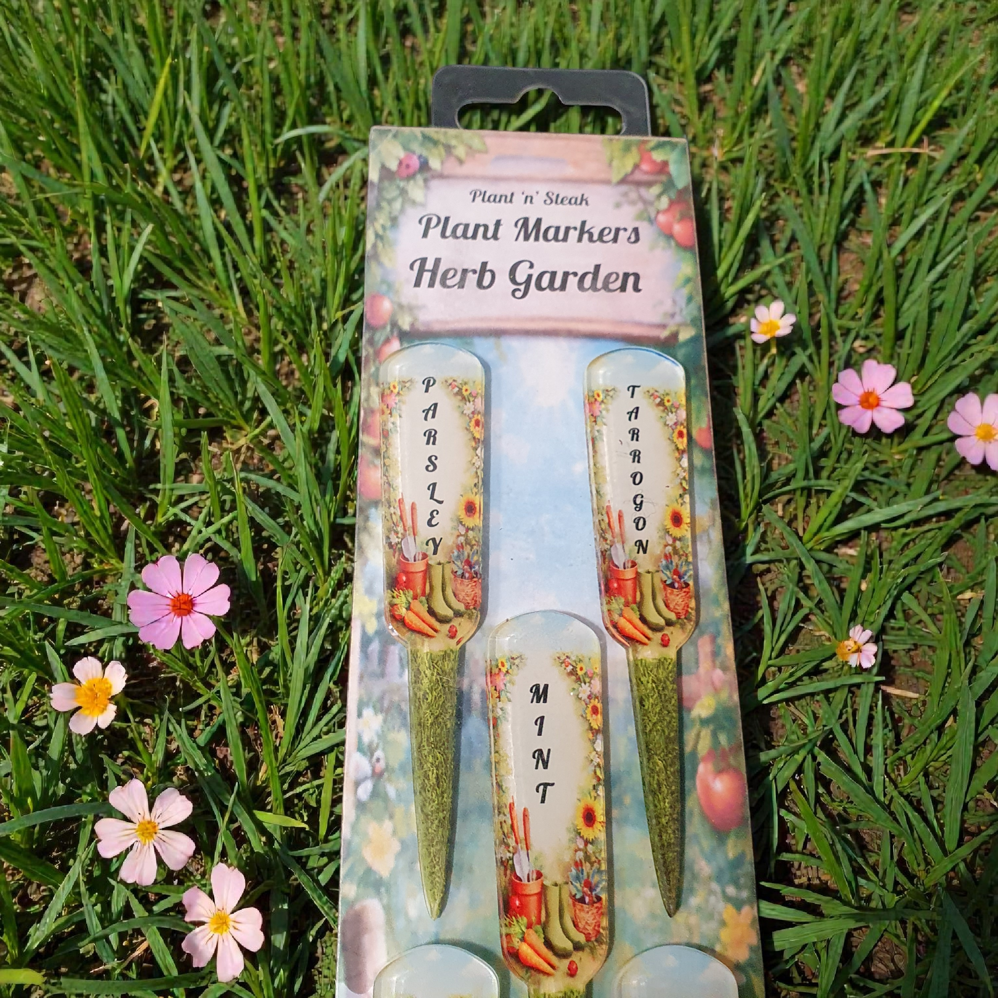 Front view of Plant ‘n’ Steak Herb Garden plant markers set on grass, featuring Parsley, Tarragon and Mint labels in illustrated packaging surrounded by flowers.