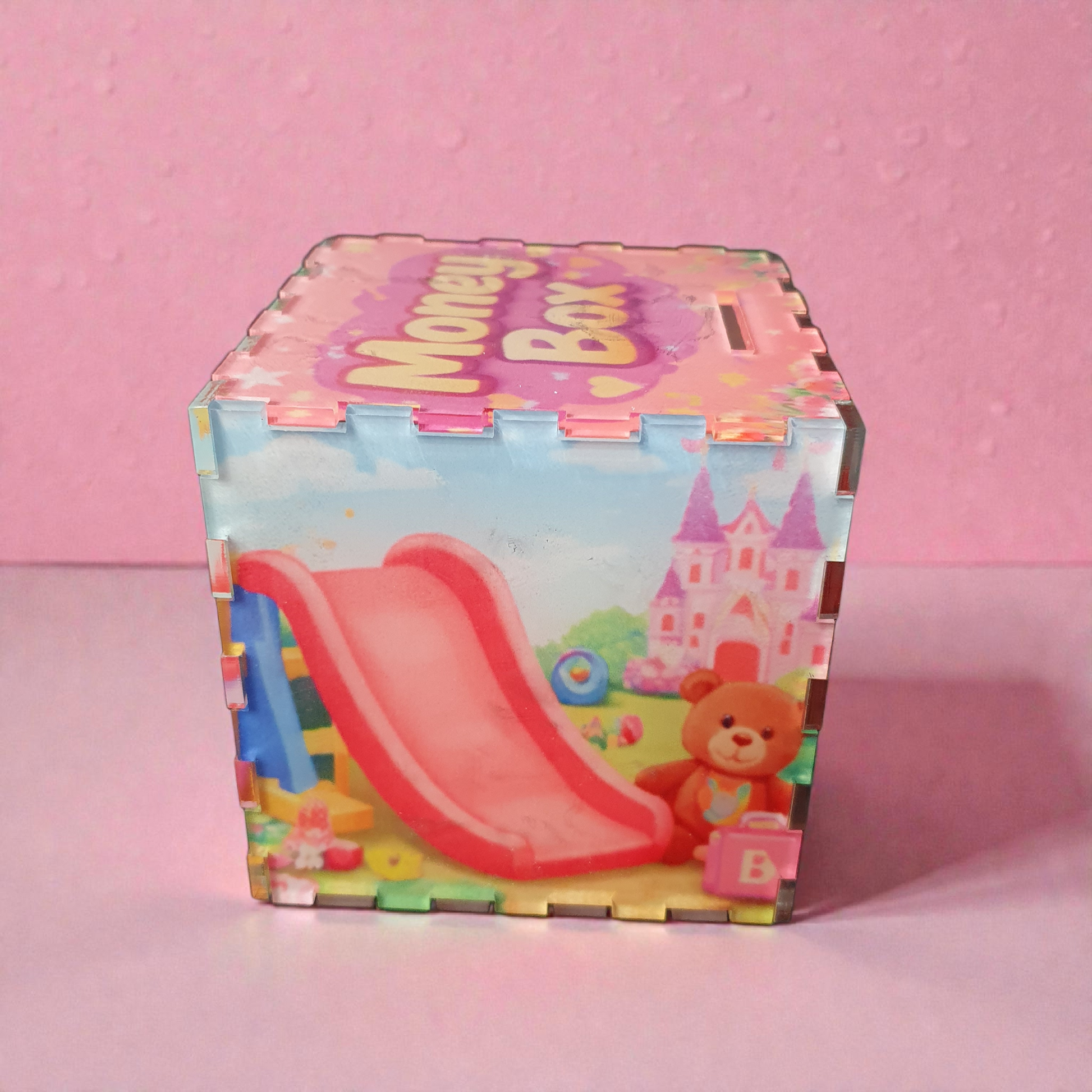 Side panel of pink children’s savings box showing playground slide, teddy bear and castle artwork.