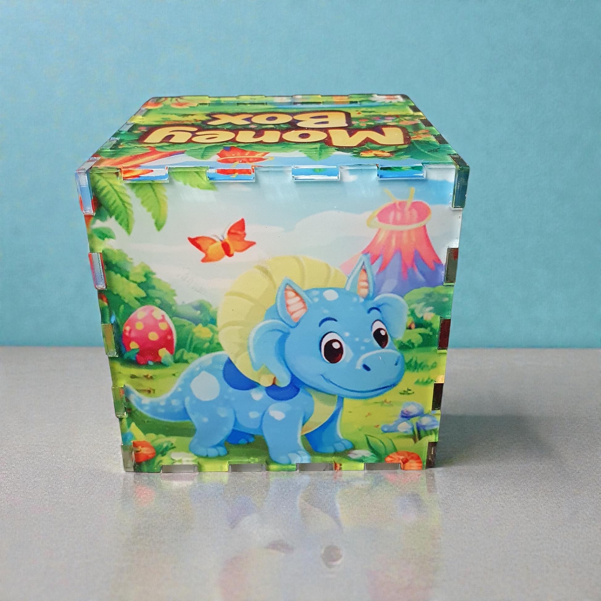 Blue triceratops style dinosaur money box with volcano and forest background, cube shaped kids coin bank.