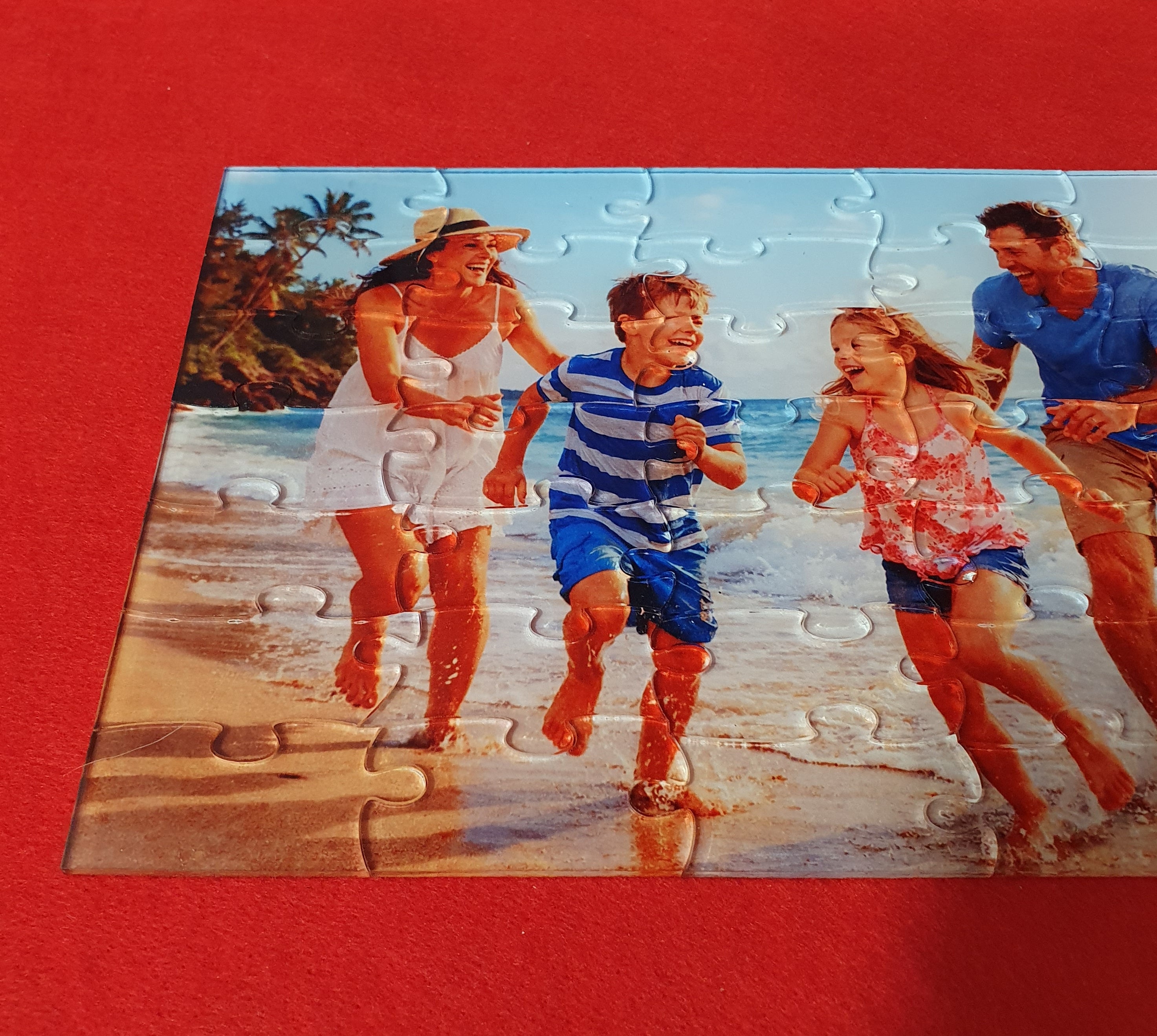 A completed jigsaw puzzle showing a family of four running and laughing on a beach, placed on a solid red background.