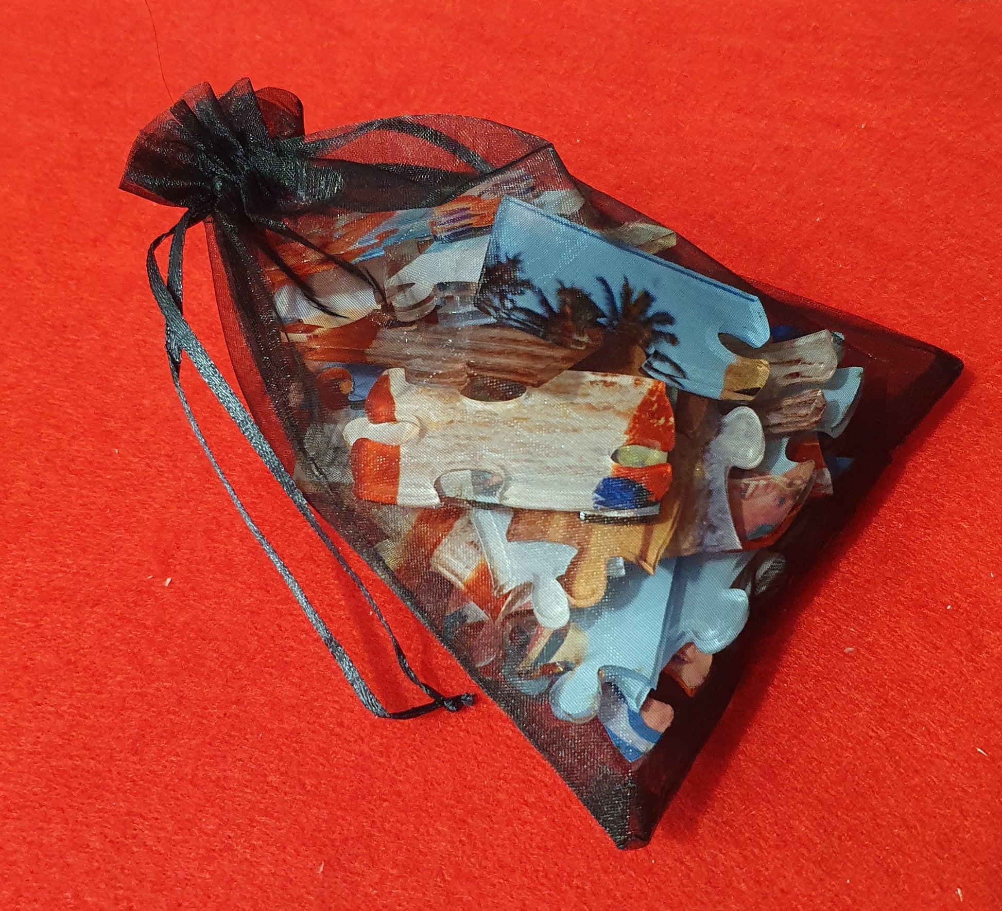 A small black sheer organza drawstring pouch filled with assorted jigsaw puzzle pieces featuring a beach scene, set against a solid red background.