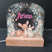 A personalized LED night light with a wooden side support, featuring an illustration of two baby deer, flowers, and the name 'Ariana' in pink script on a dark background.