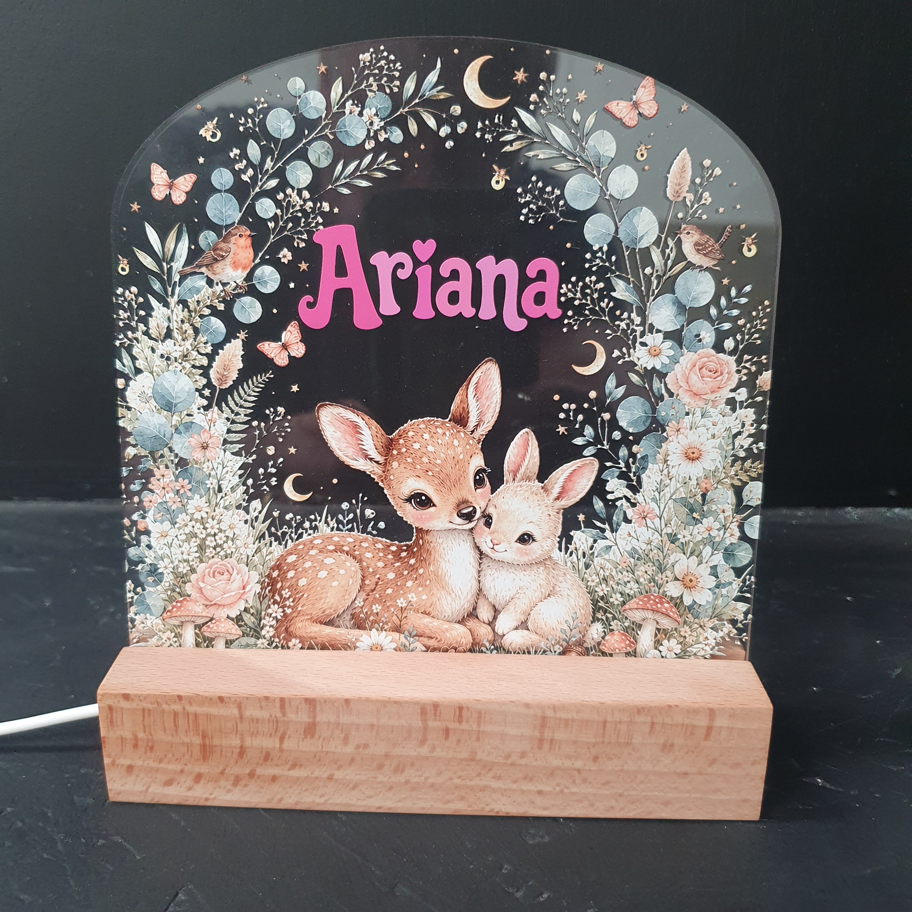 A personalized LED night light with a wooden side support, featuring an illustration of two baby deer, flowers, and the name 'Ariana' in pink script on a dark background.