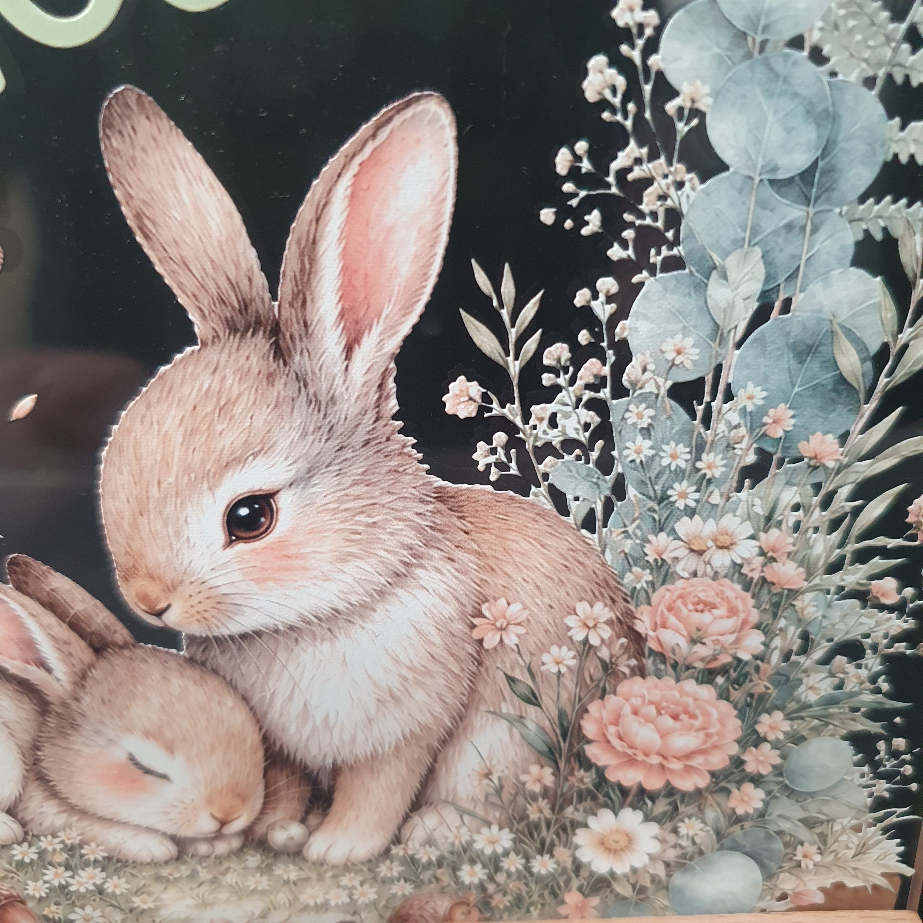 A close-up of a decorative surface featuring a detailed illustration of two rabbits nestled among soft pink flowers and eucalyptus leaves on a dark background.