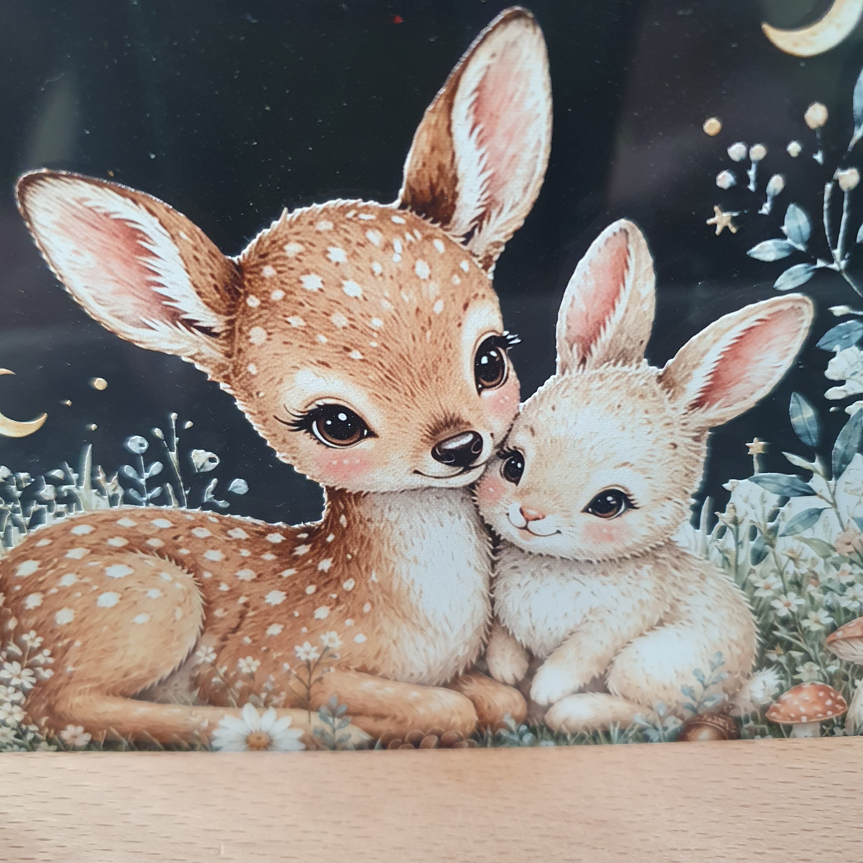 A close-up of a decorative surface featuring an illustrated scene of two spotted deer, one fawn and one young rabbit, with floral and crescent-moon details on a dark background. A light wood edge is visible along the right side.