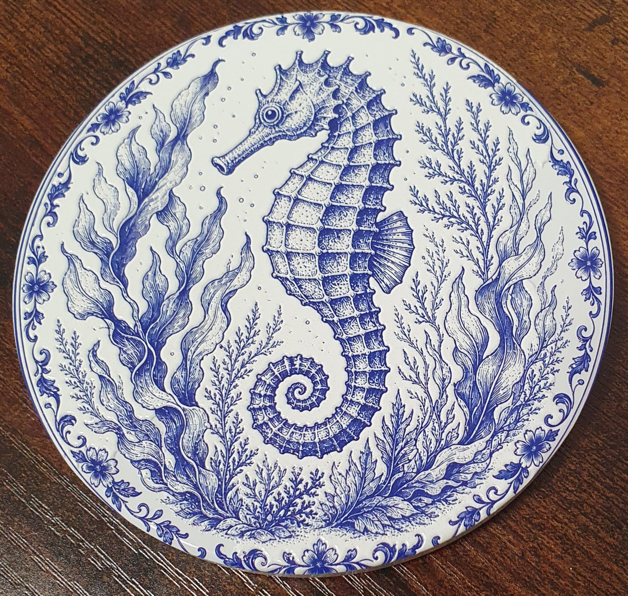A round white ceramic coaster with a detailed blue seahorse and sea plant pattern, resting on a dark wooden surface.