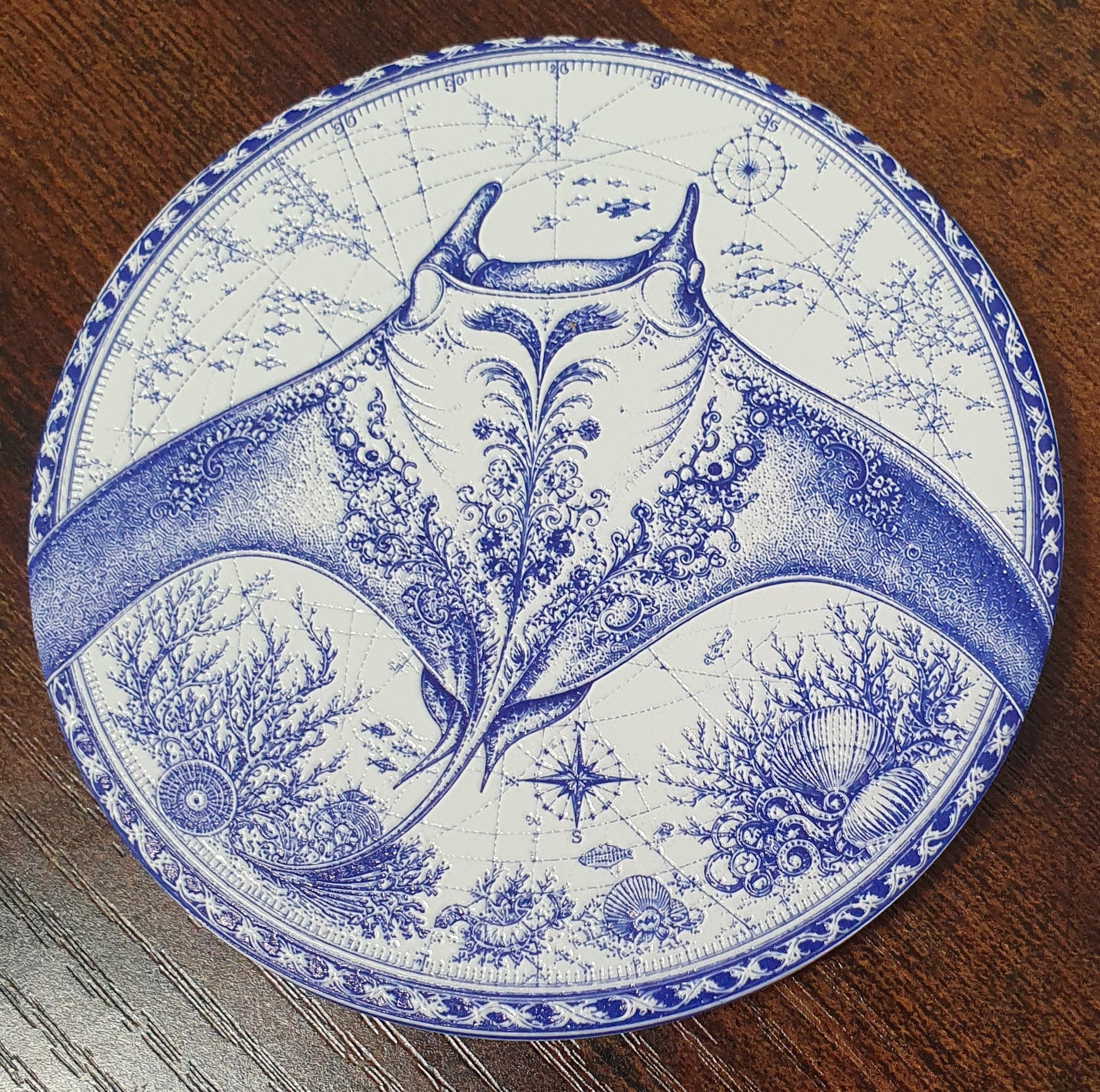 A round coastert or decorative coaster with a blue and white blue-and-white print of a manta ray and marine life, placed on a dark wood surface.