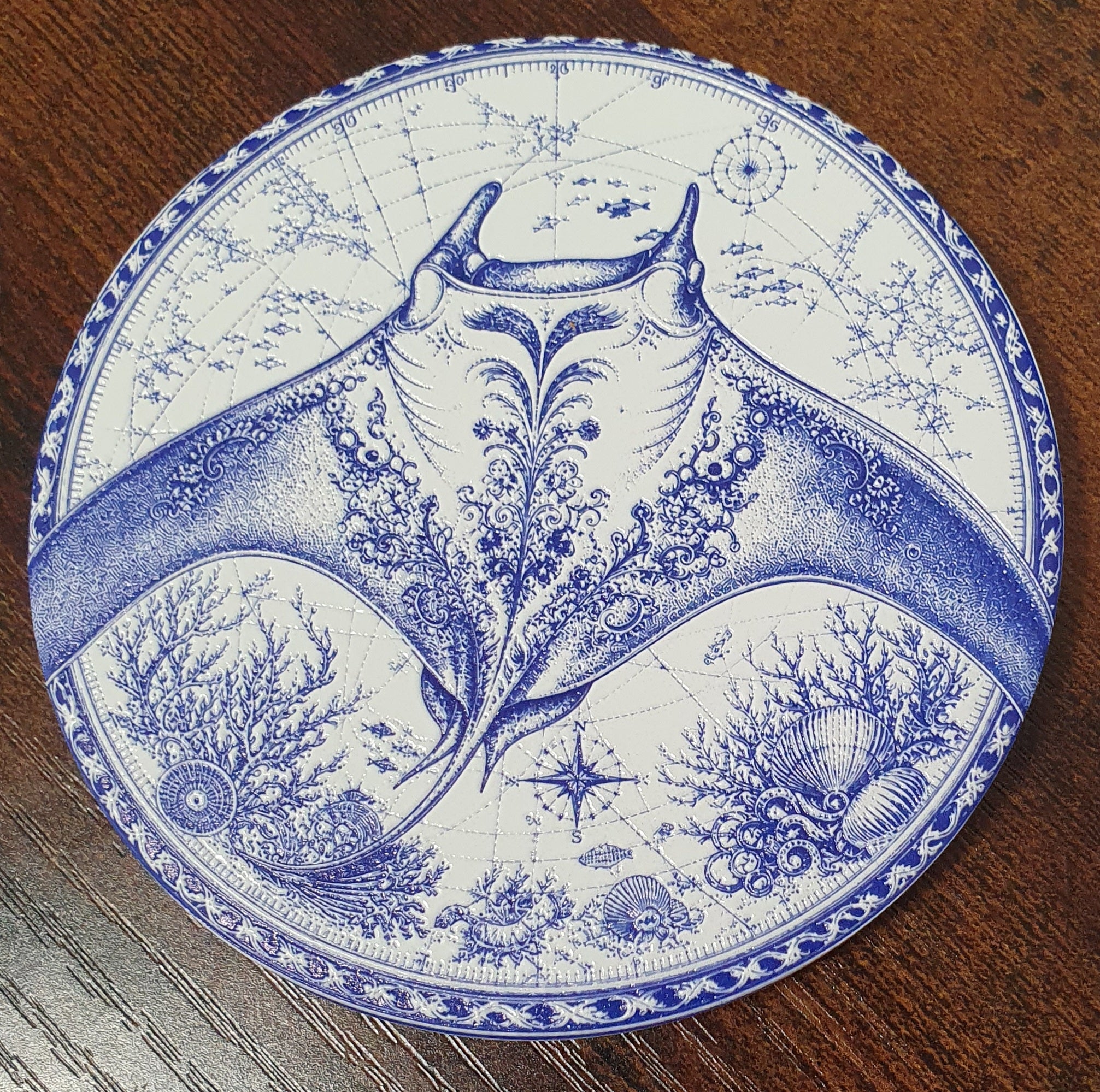 A round coastert or decorative coaster with a blue and white blue-and-white print of a manta ray and marine life, placed on a dark wood surface.
