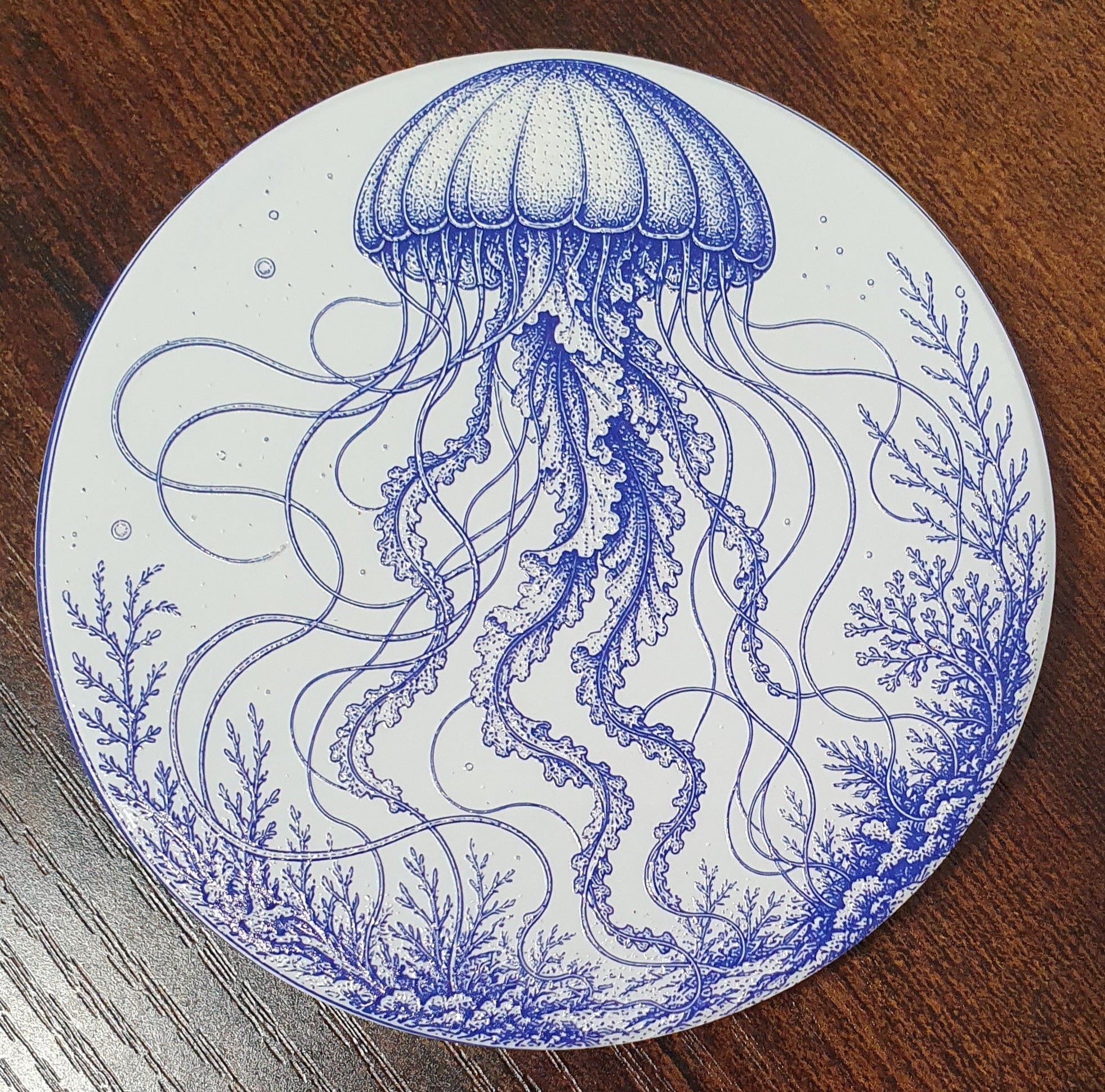 A round white coaster featuring a detailed blue line art illustration of a jellyfish and marine plants, photographed on a dark wood surface.