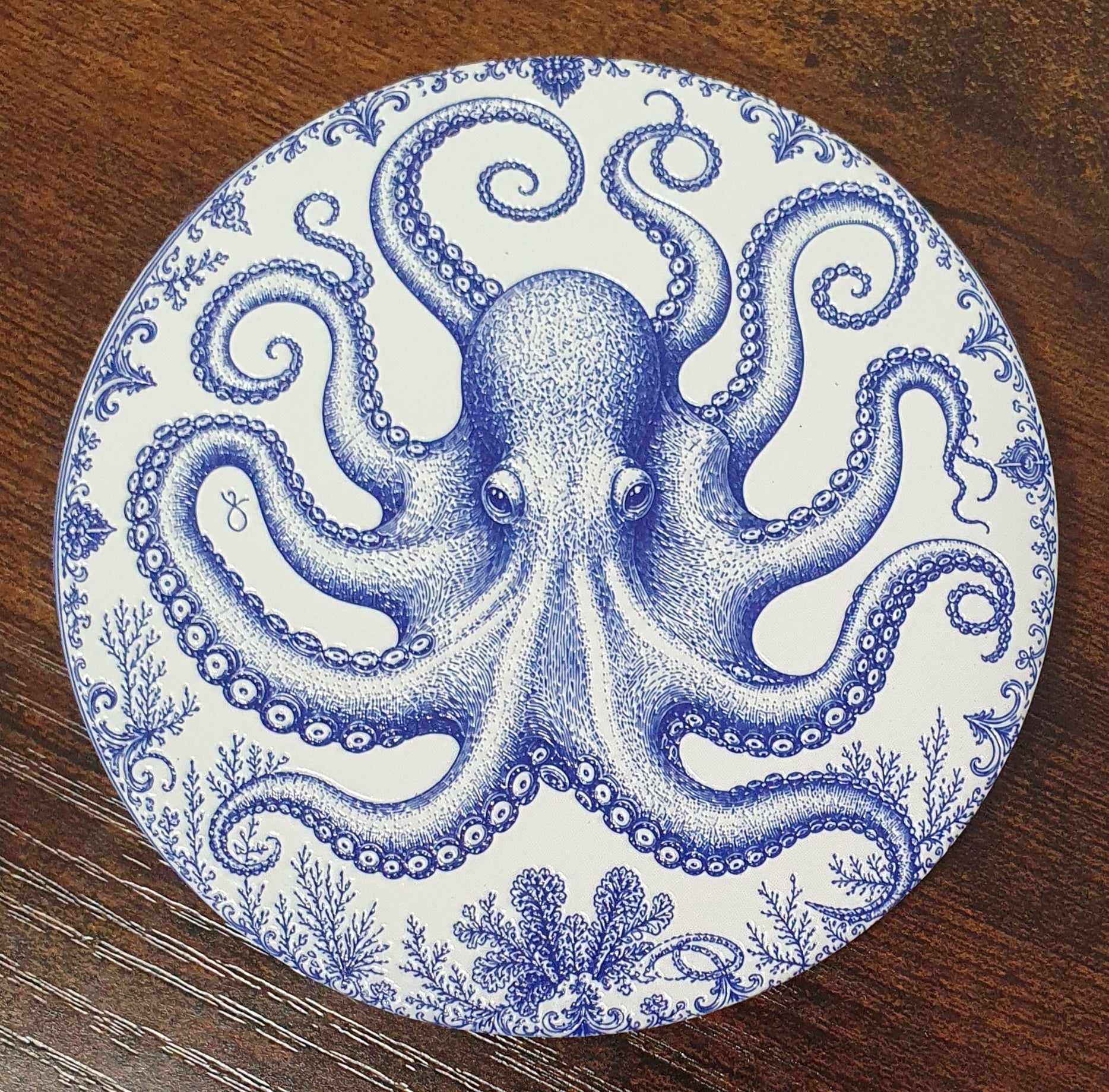 A round coaster featuring a detailed blue octopus illustration and small sea plant motifs, photographed on a dark wood surface.