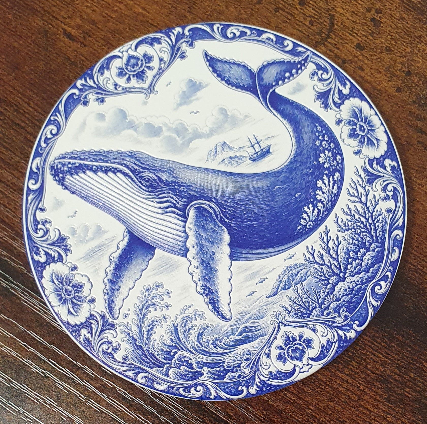 A round blue and white ceramic coaster featuring a detailed illustration of a blue whale in an ocean scene, with decorative floral and scroll borders, placed on a dark wooden surface.