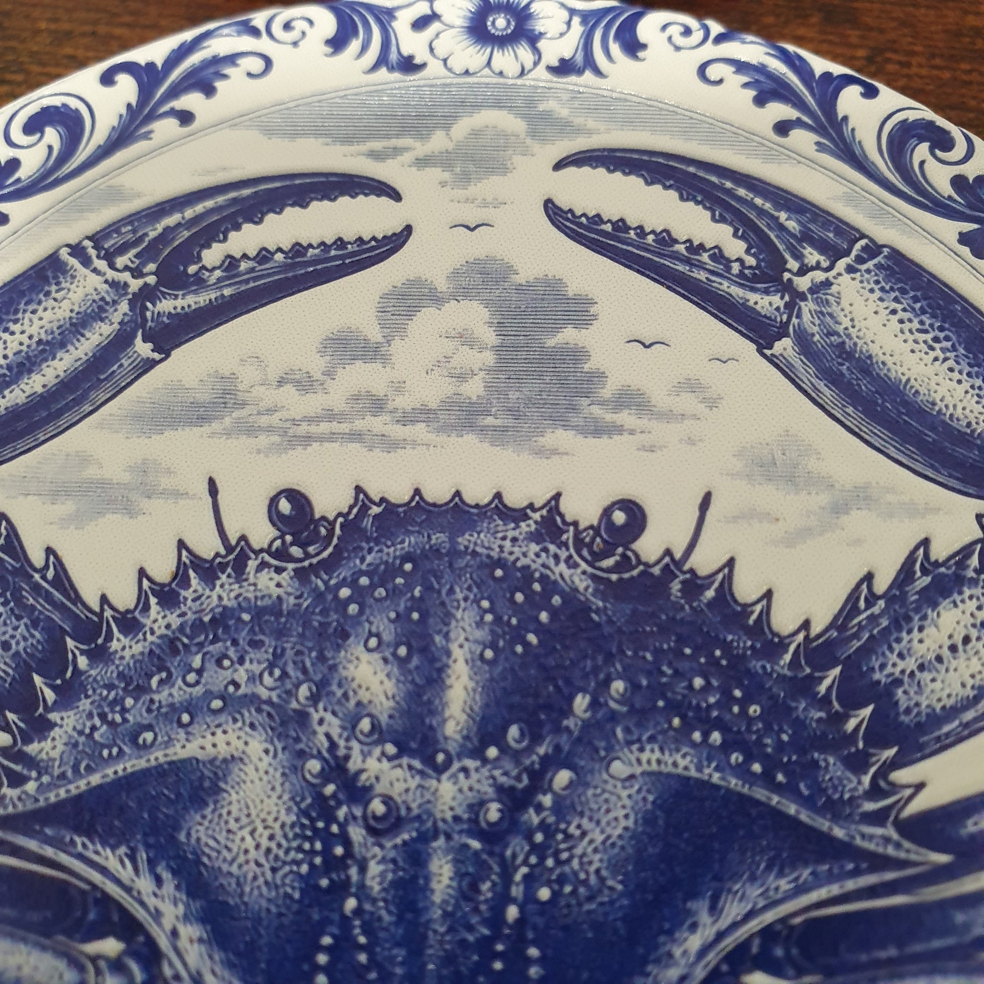 Close-up of a blue and white ceramic coaster featuring a detailed crab illustration and ornate floral scrollwork around the rim.