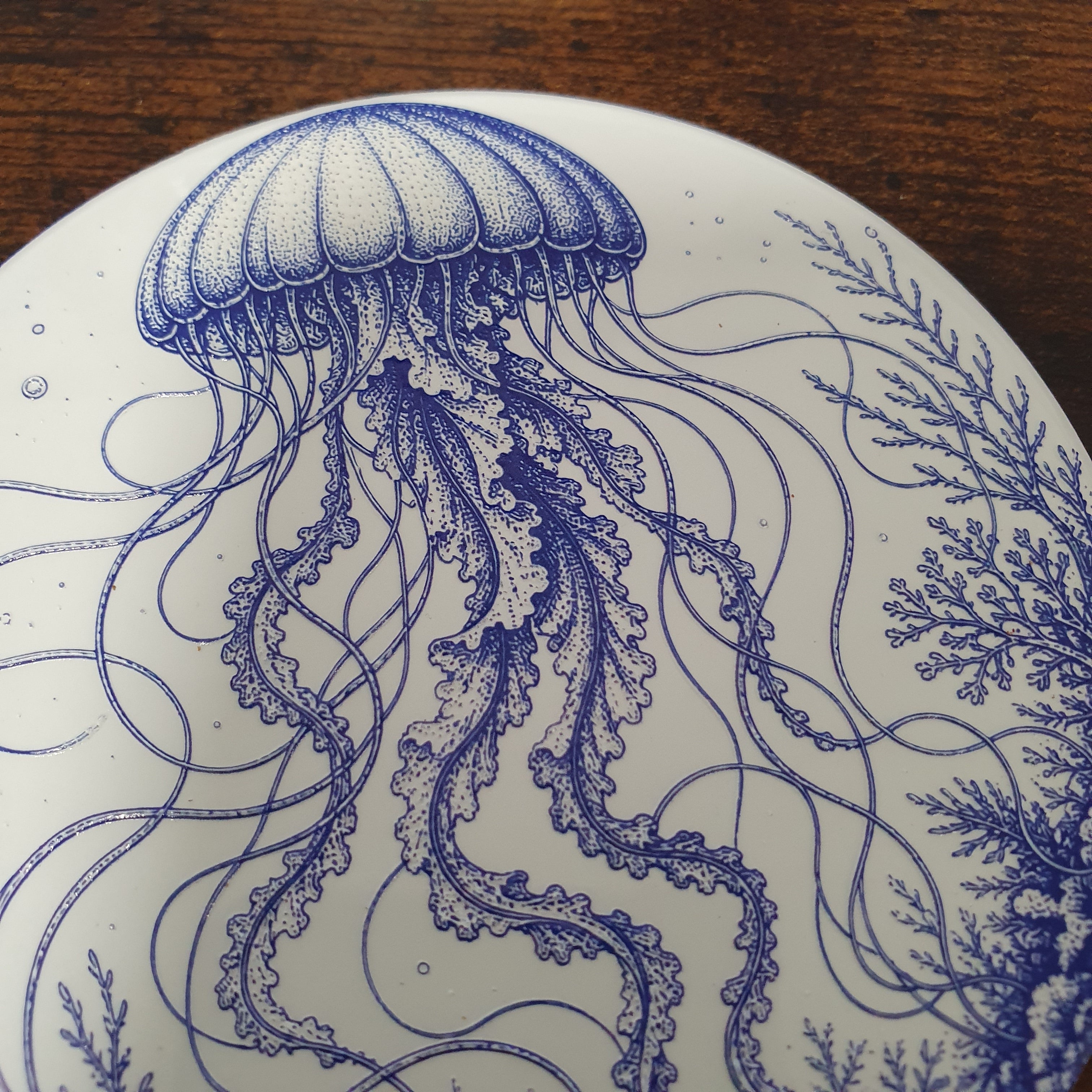 A close-up of a white coaster featuring a detailed blue illustration of a jellyfish and marine plants, set against a dark wooden background.