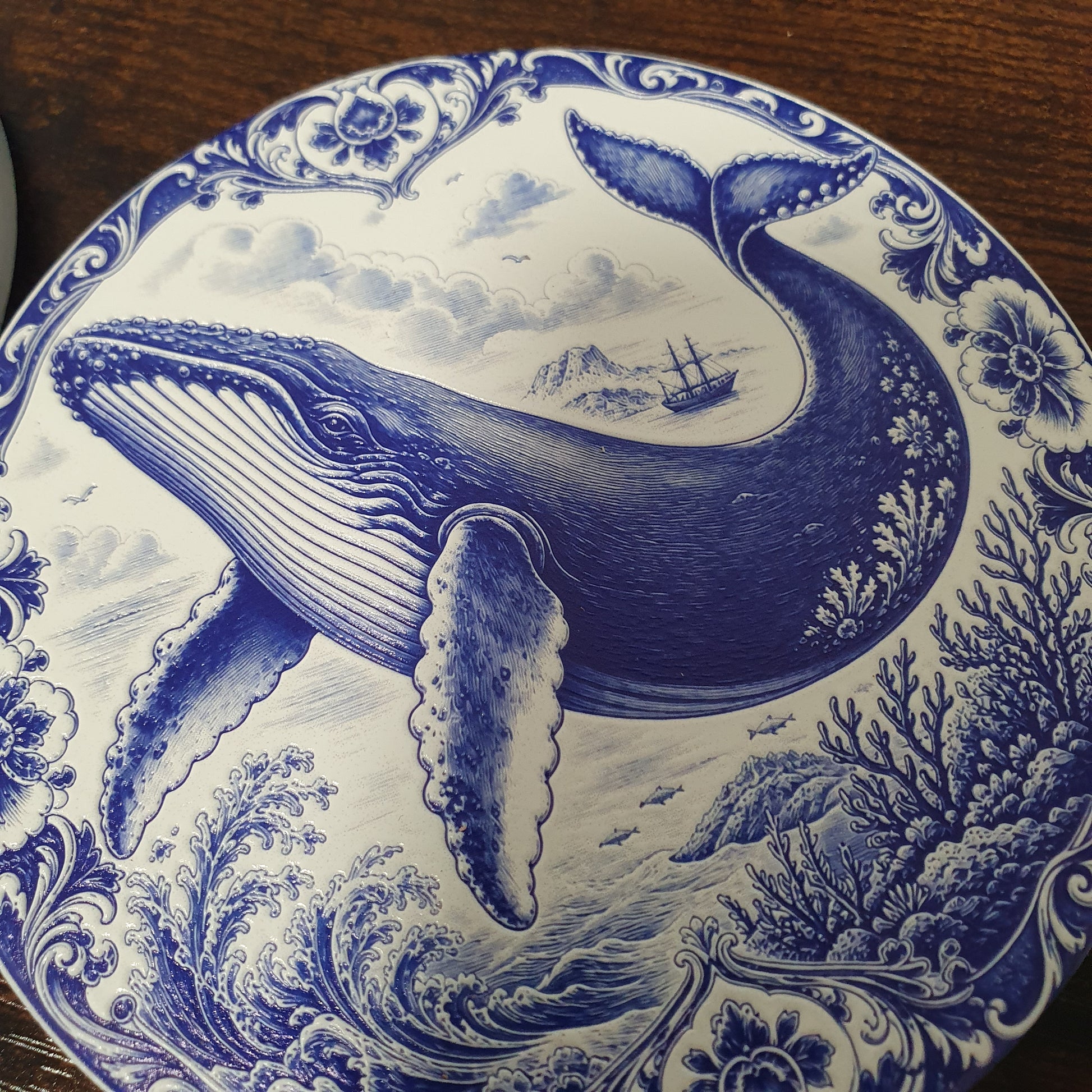 A round ceramic coaster featuring a detailed blue and white illustration of a whale, waves, coral, and a ship, photographed close-up on a dark wooden surface.