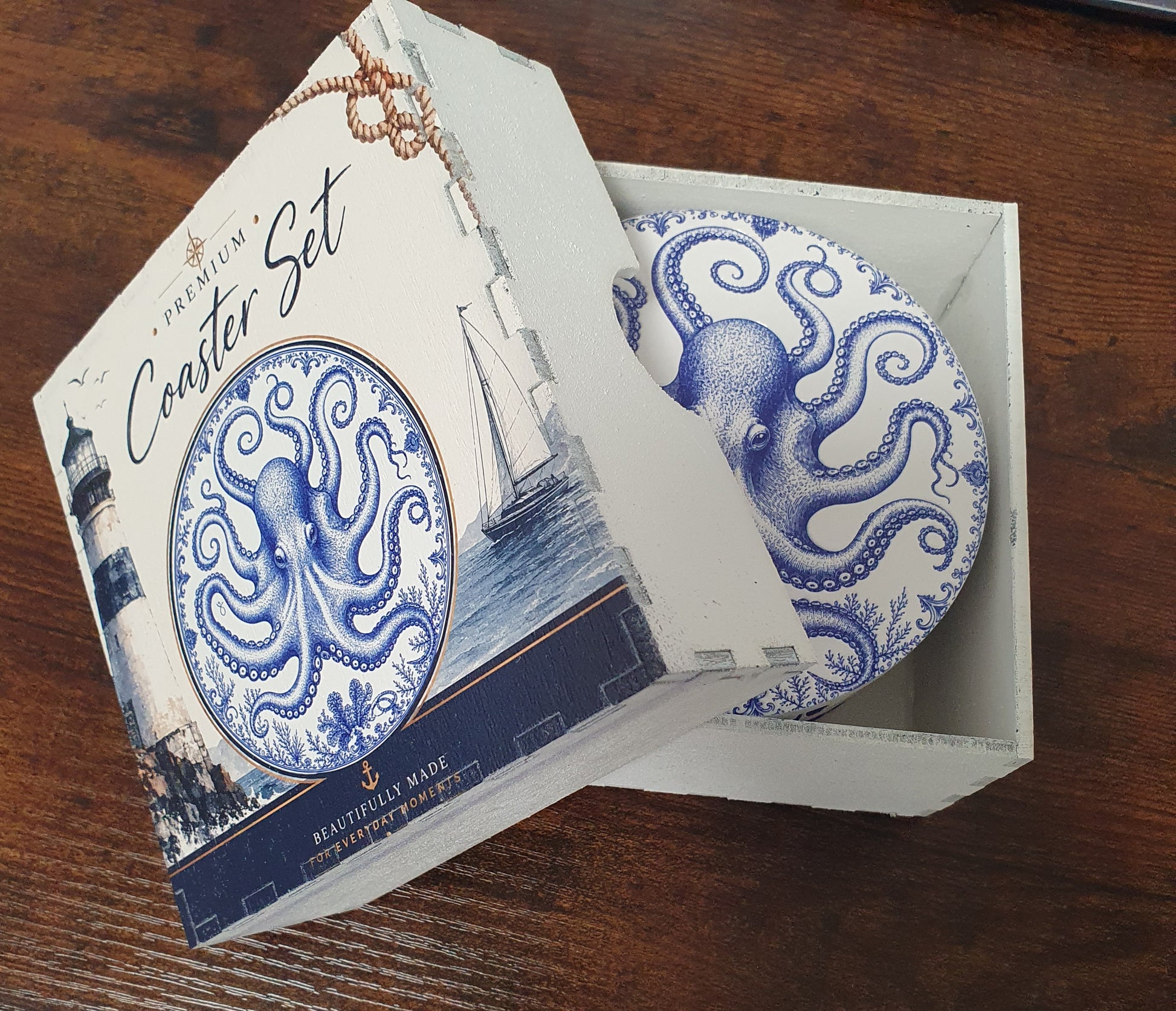 A premium coaster set in its decorative packaging, featuring a round blue and white octopus coaster visible inside a white box with a lighthouse illustration on the lid, placed on a dark wooden table.