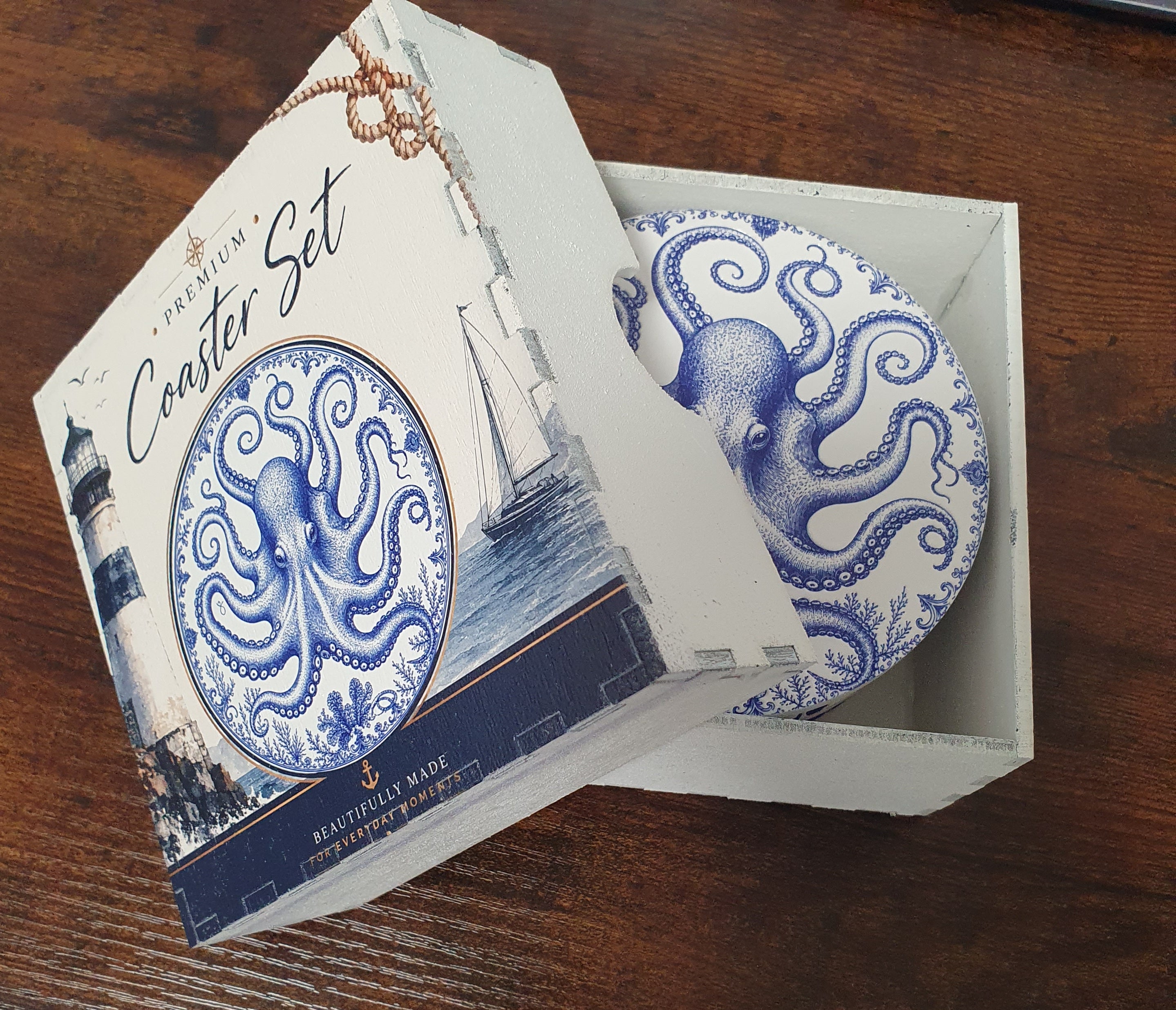 A premium coaster set in its decorative packaging, featuring a round blue and white octopus coaster visible inside a white box with a lighthouse illustration on the lid, placed on a dark wooden table.