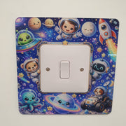A square light switch plate cover featuring a colorful cartoon space theme with astronauts, planets, stars, and a rocket, shown installed over a white single-toggle switch on a plain light background.
