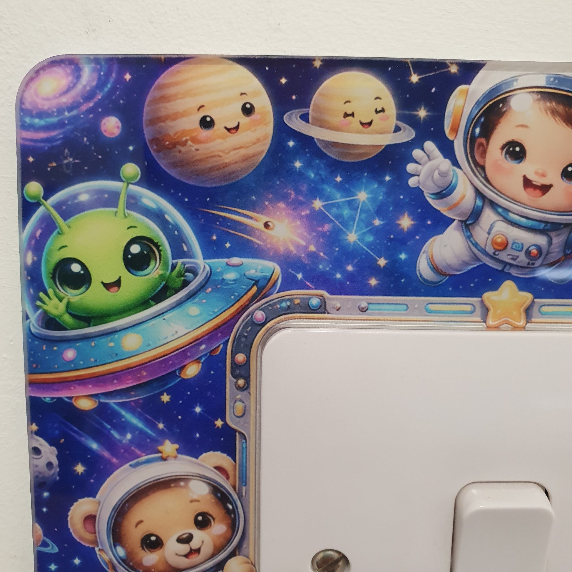 A close-up of a white light switch plate decorated with a colorful cartoon space design, featuring planets, a green alien in a UFO, and smiling astronauts including a bear astronaut.