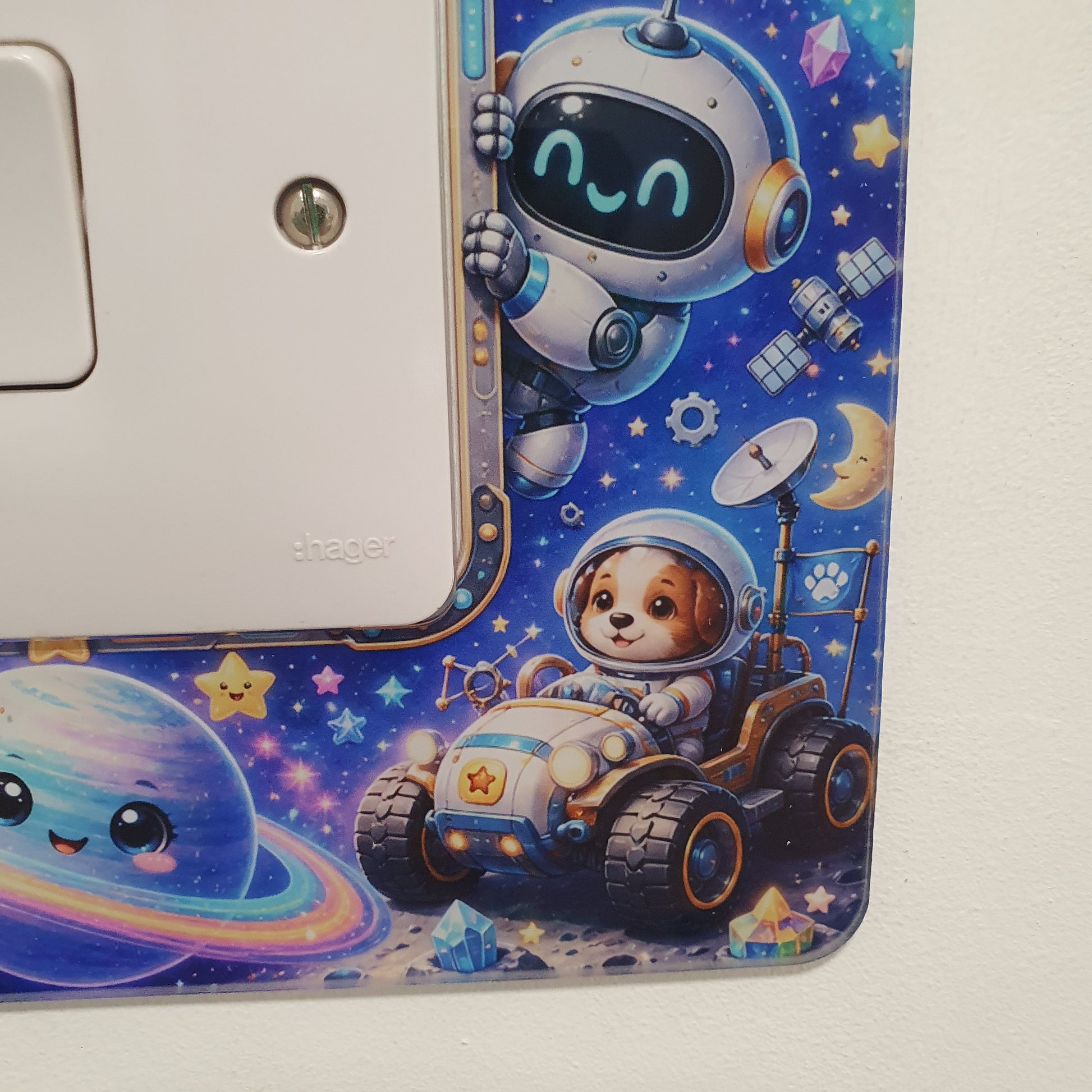 A close-up of a white electrical outlet cover plate decorated with a colorful cartoon space theme, featuring astronauts, a rover, stars, and planets on a blue starry background.