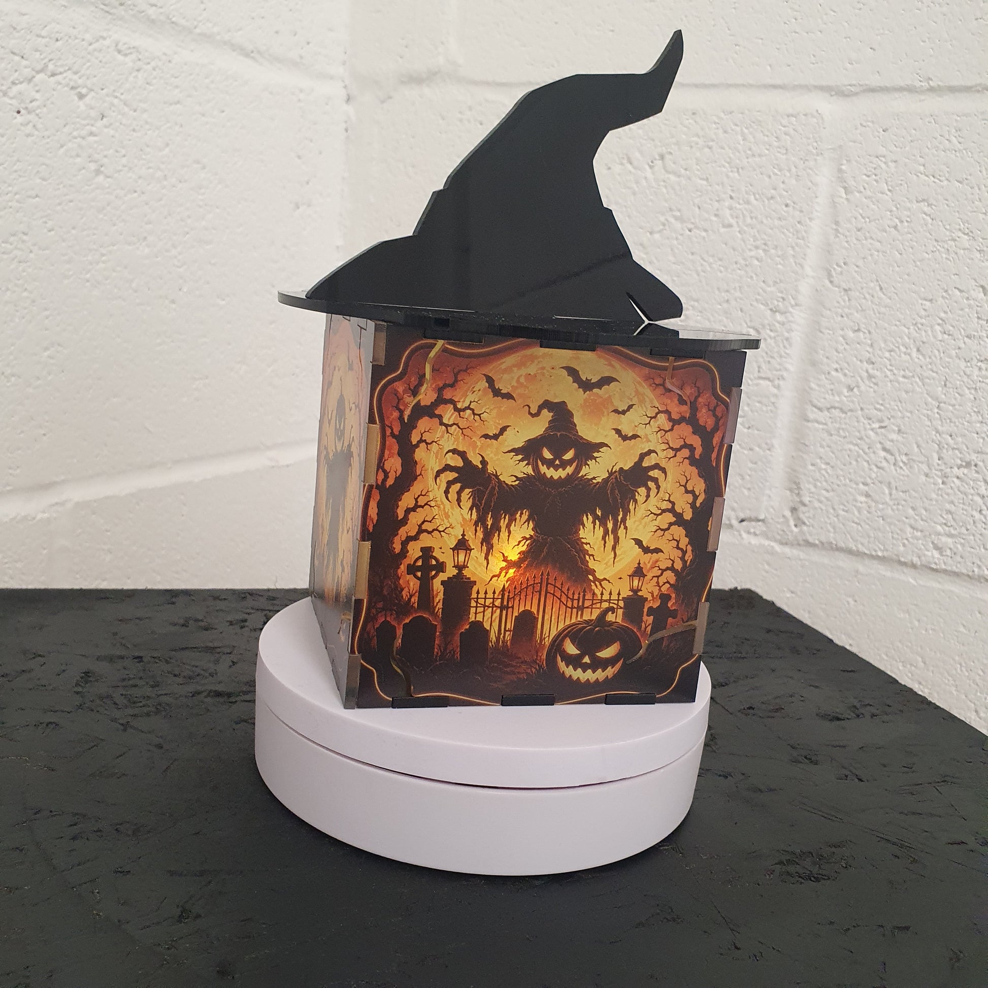 A Halloween-themed decorative box with a black witch hat on top, featuring a glowing orange and yellow haunted house scene on the front, displayed on a white circular base against a white brick wall.