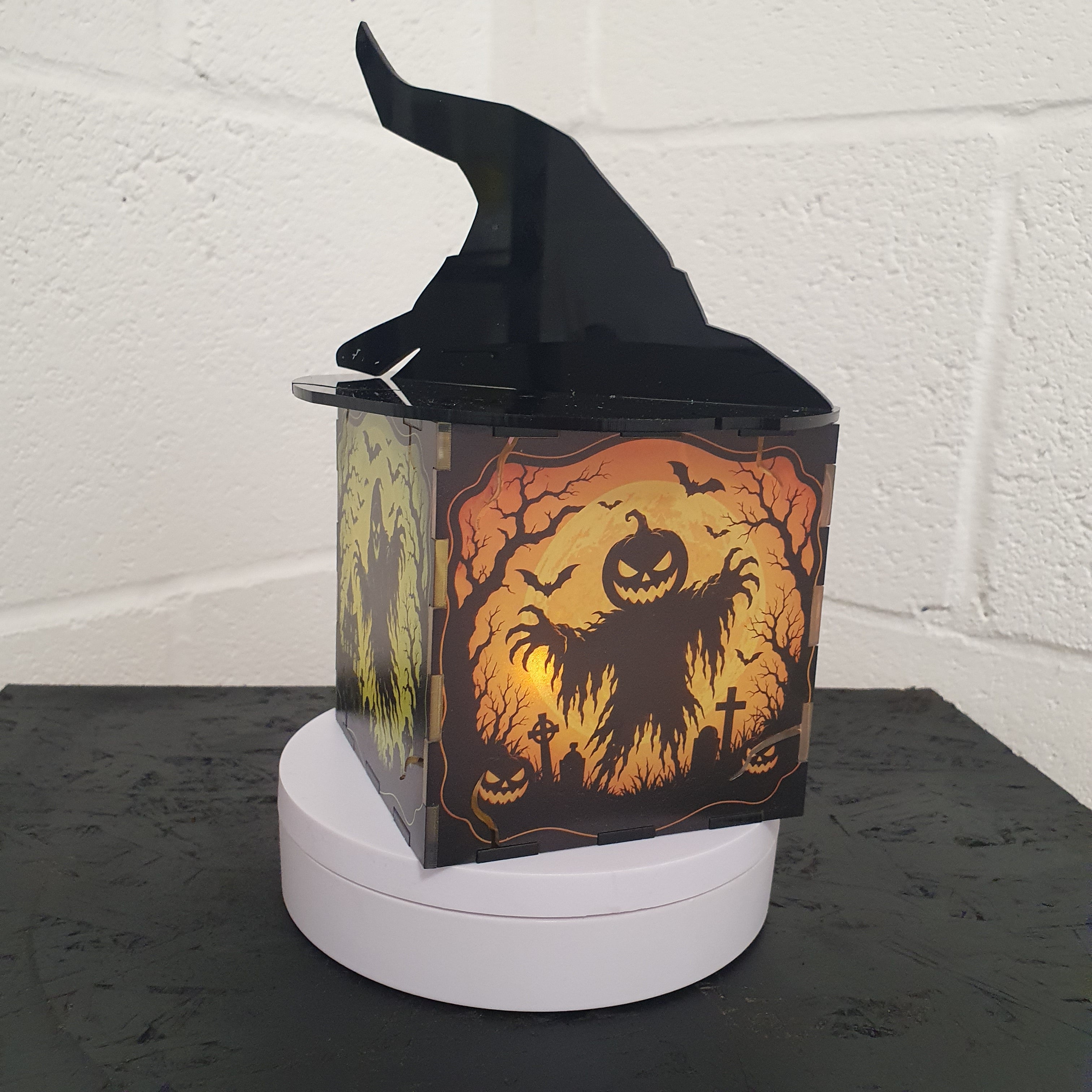A Halloween-themed decorative box with a black witch hat lid, featuring a spooky orange and black ghost illustration on the front, displayed on a white circular base against a white brick wall.