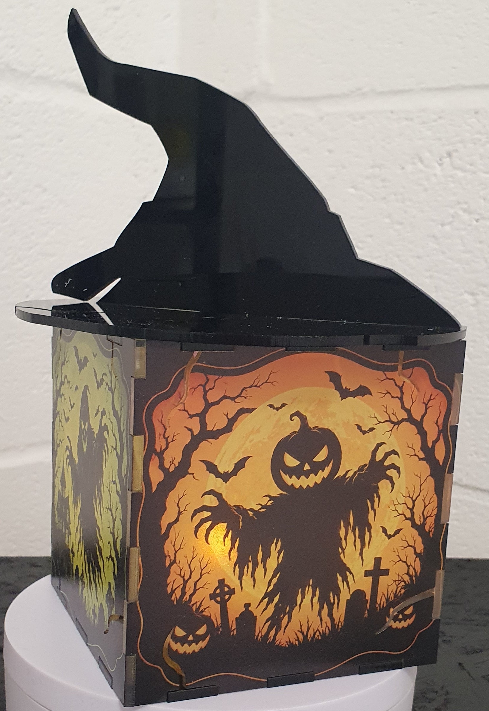 A Halloween-themed decorative box with a black witch hat on top and a spooky orange and black haunted-house illustration on the front, displayed on a white pedestal against a white wall.