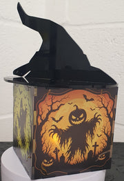 A Halloween-themed decorative box with a black witch hat on top and a spooky orange and black haunted-house illustration on the front, displayed on a white pedestal against a white wall.