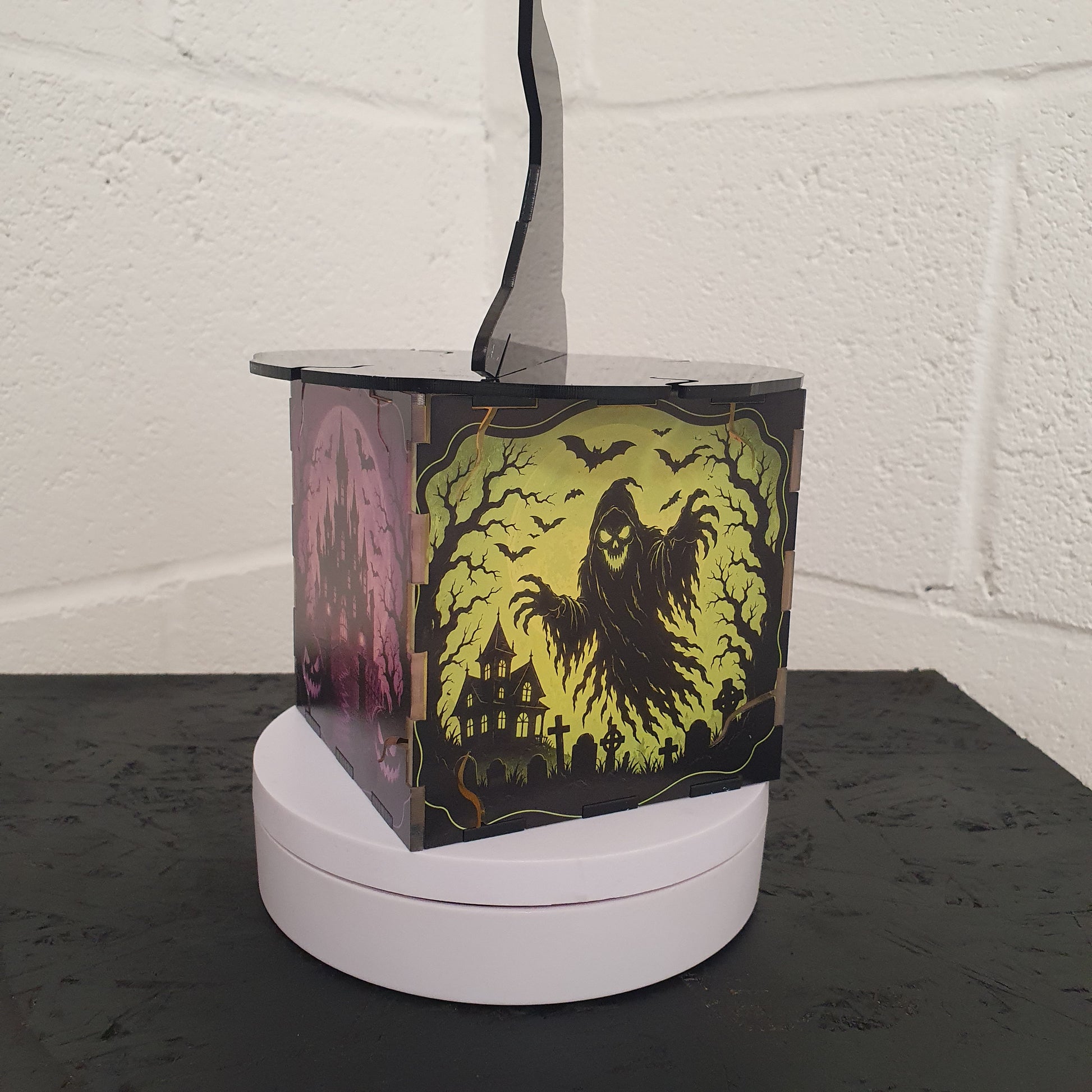 A square Halloween-themed table lamp with a rotating display base, featuring a black and yellow ghostly figure design on the side panels, set against a white brick wall background.