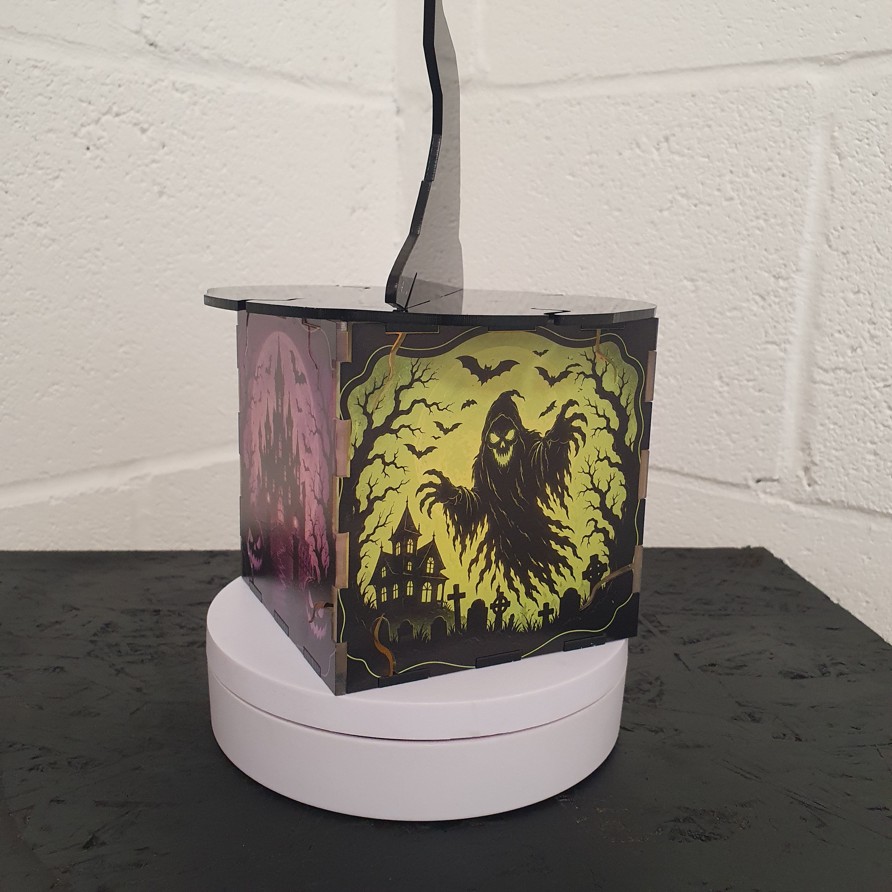 A square Halloween-themed table lamp with a rotating display base, featuring a black and yellow ghostly figure design on the side panels, set against a white brick wall background.