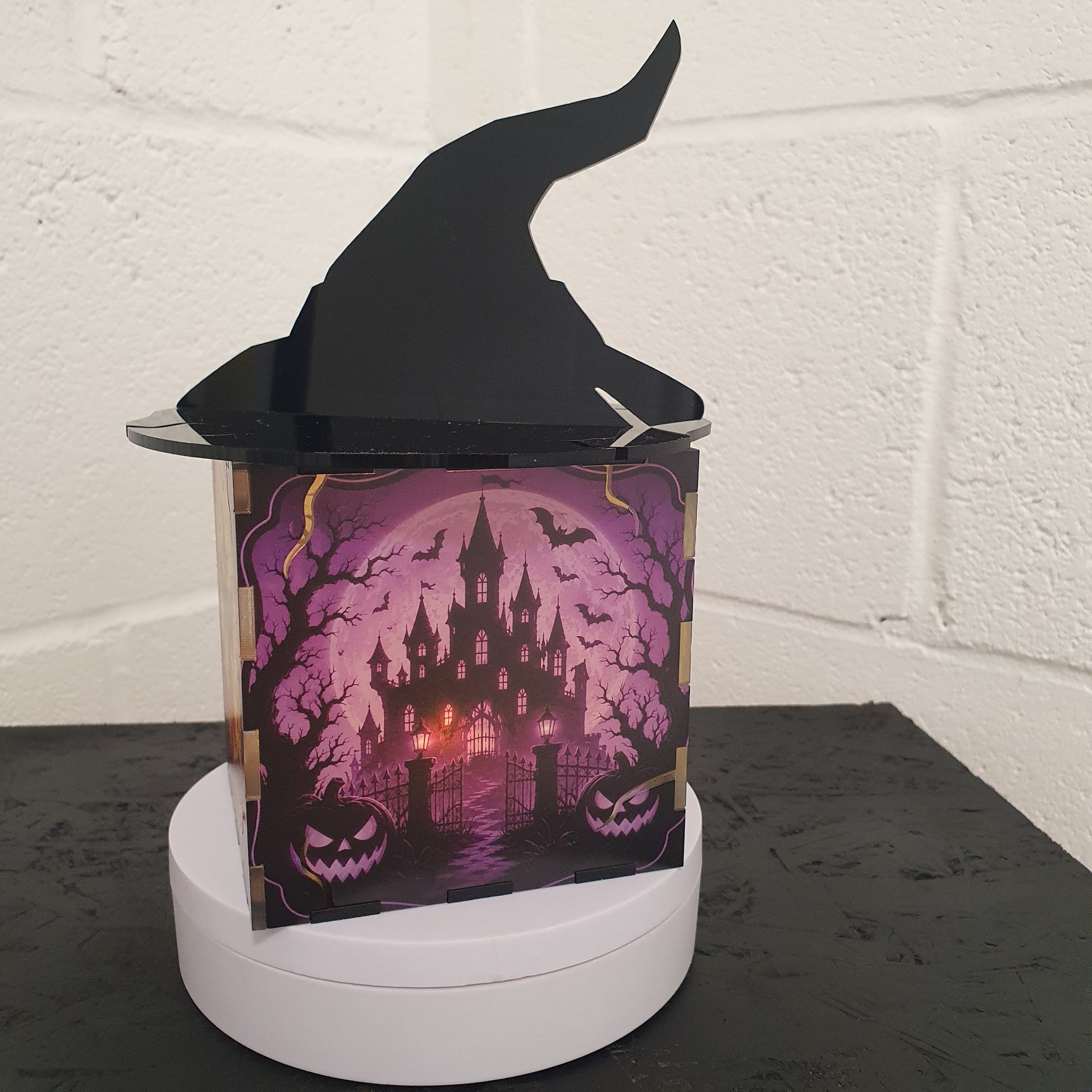 A Halloween-themed decorative box featuring a black witch hat and a purple haunted castle illustration, displayed on a white circular base against a white brick wall.