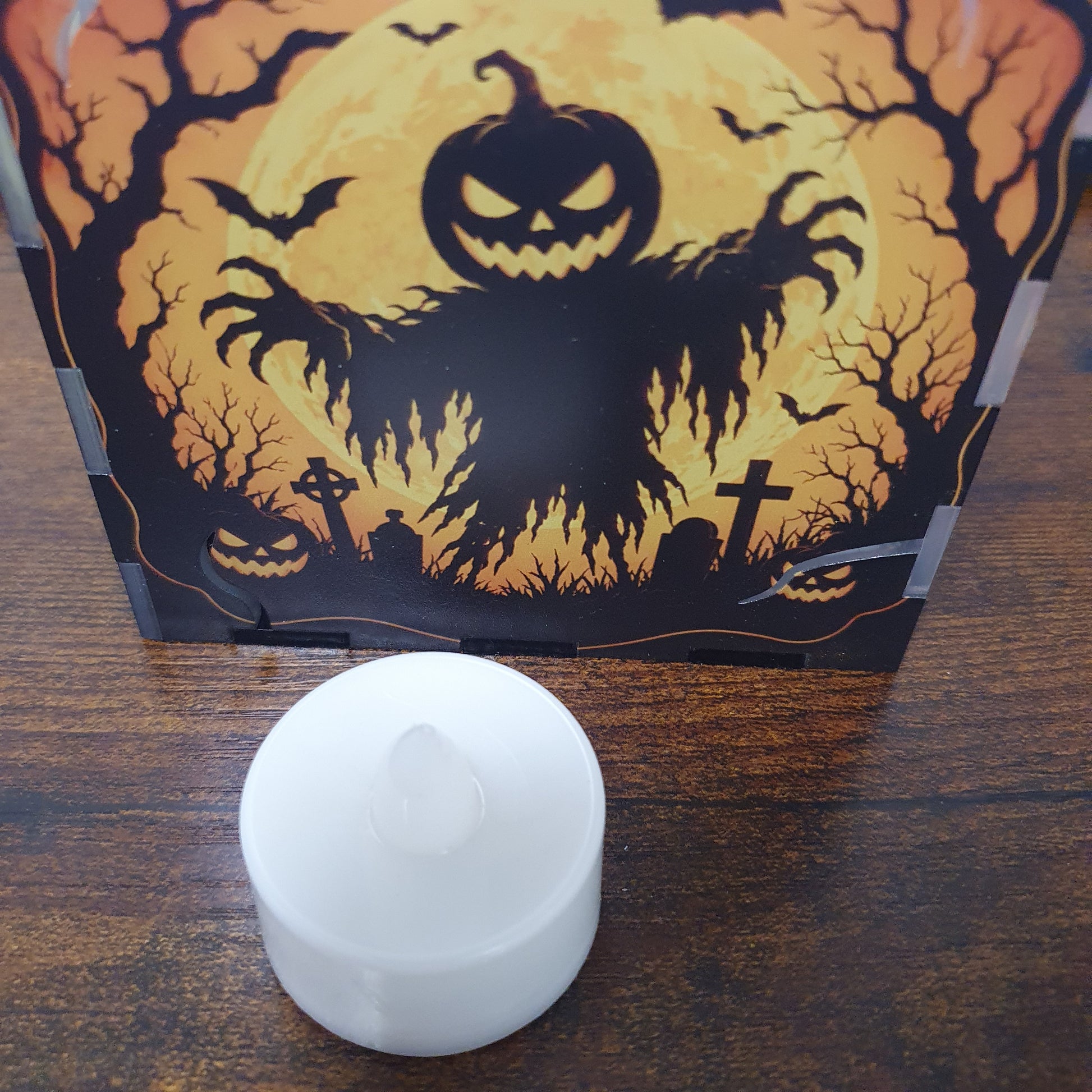 A small white LED tea light candle sits in front of its Halloween-themed packaging box, which features a spooky jack-o'-lantern, bats, and a full moon design on a dark wooden tabletop.