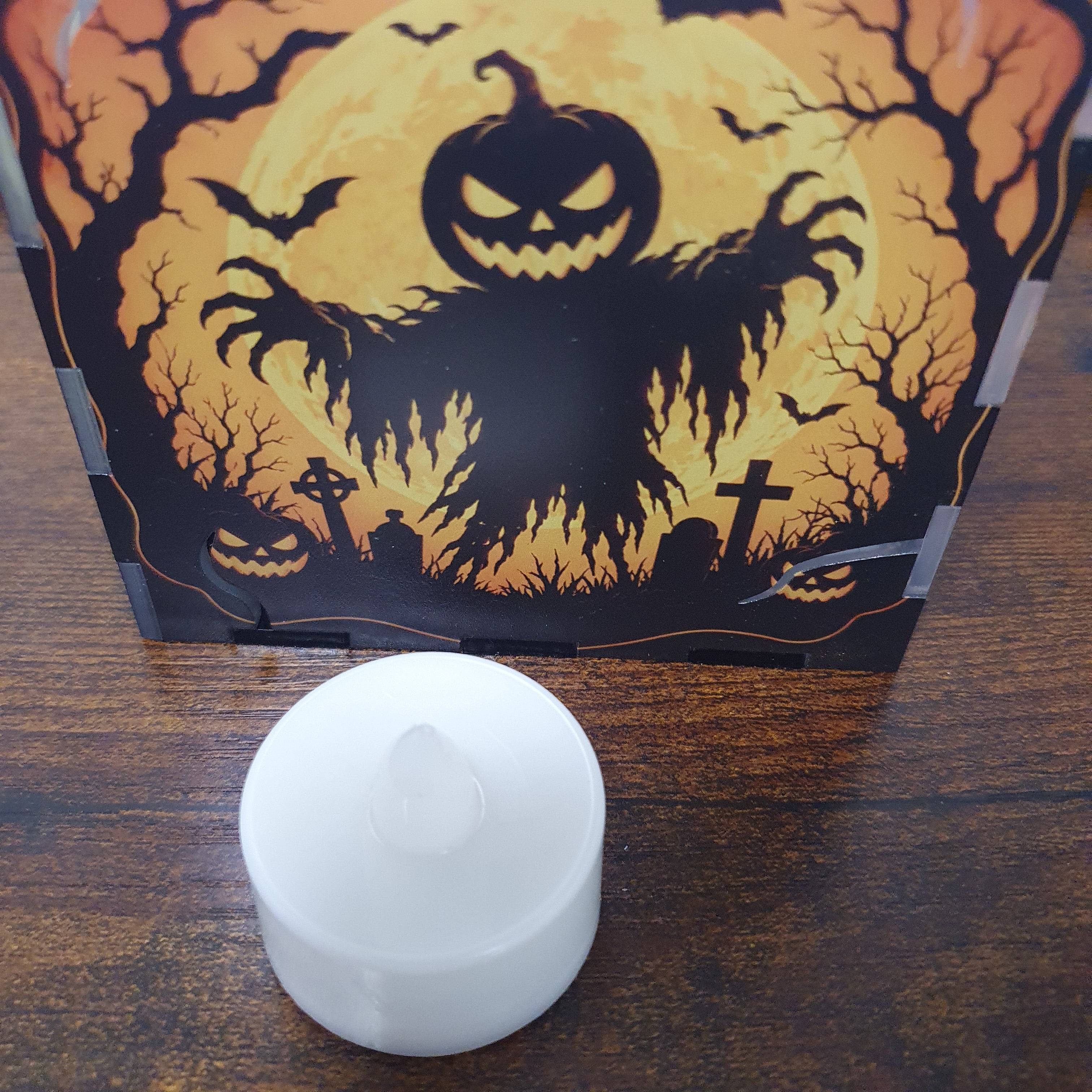 A small white LED tea light candle sits in front of its Halloween-themed packaging box, which features a spooky jack-o'-lantern, bats, and a full moon design on a dark wooden tabletop.