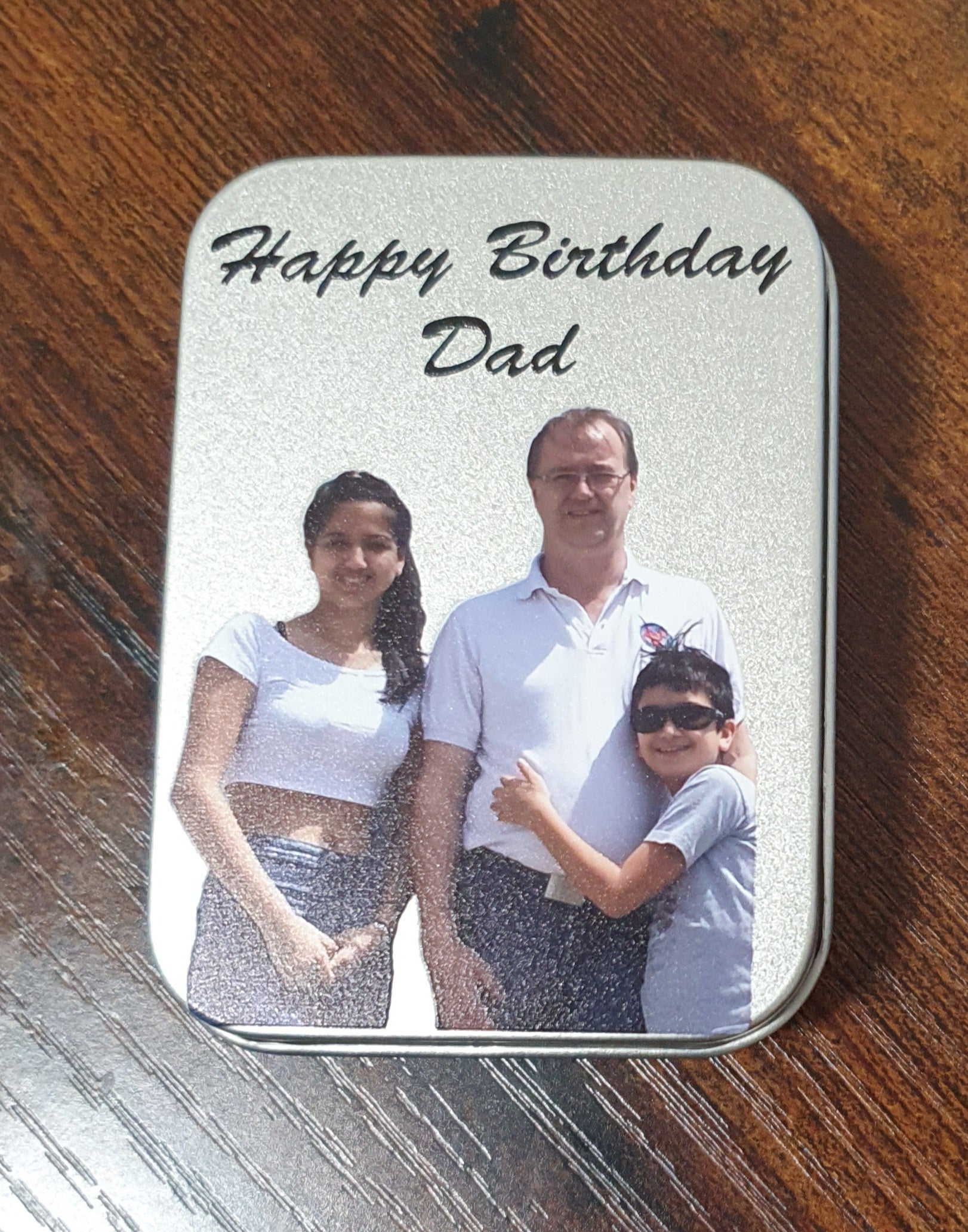 Personalised Lighter Gift Box. A rectangular metal tin with rounded corners sits on a wooden table. The tin has a silver glittery background with the words 'Happy Birthday Dad' printed in black script at the top, and a printed family photo of a father, mother, and son in the center.