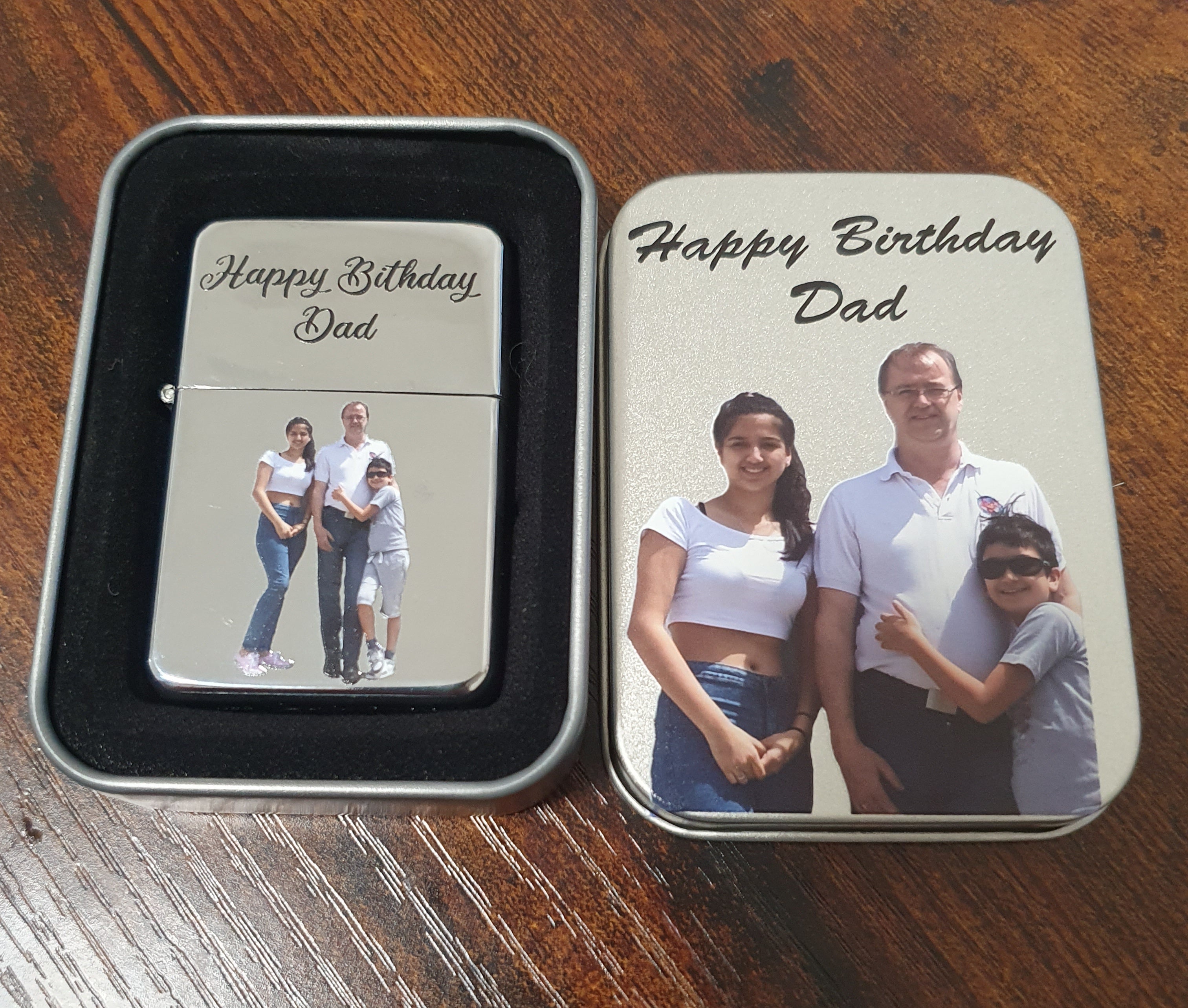 A personalized metal tin lighter and matching tin box on a wooden table. The lighter is silver with a black insert and the text 'Happy Birthday Dad' above a family photo.