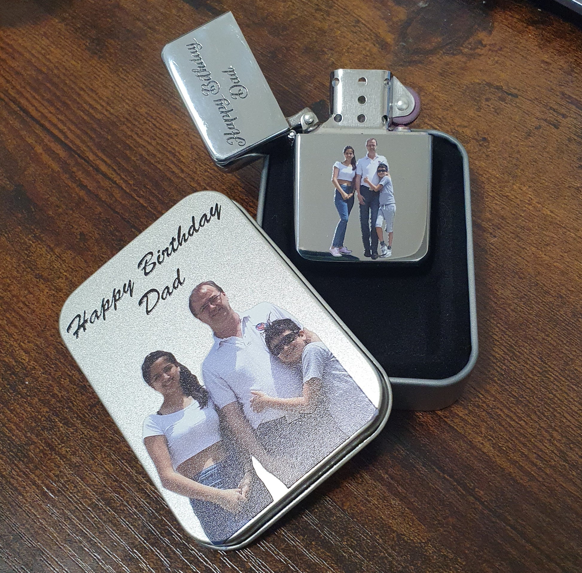 A set of custom photo metal lighter covers and a matching lighter displayed on a wooden table. The covers are engraved with 'Happy Birthday Dad' and feature a printed photo of a father and two children.