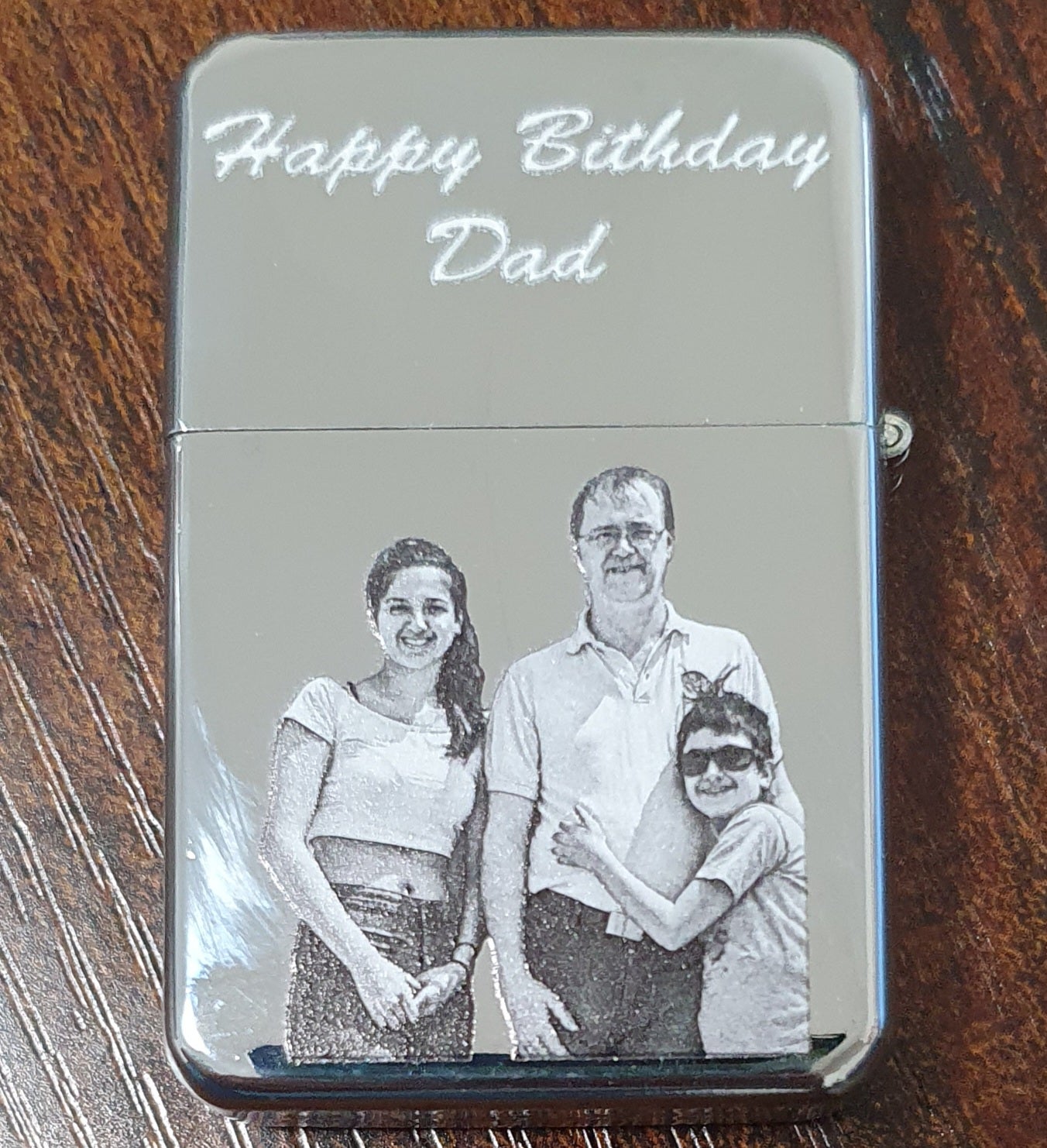 A custom-engraved chrome lighter resting on a dark wooden table. The lighter features the text 'Happy Birthday Dad' in white script at the top, and a black-and-white family portrait of a father, mother, and son engraved below.