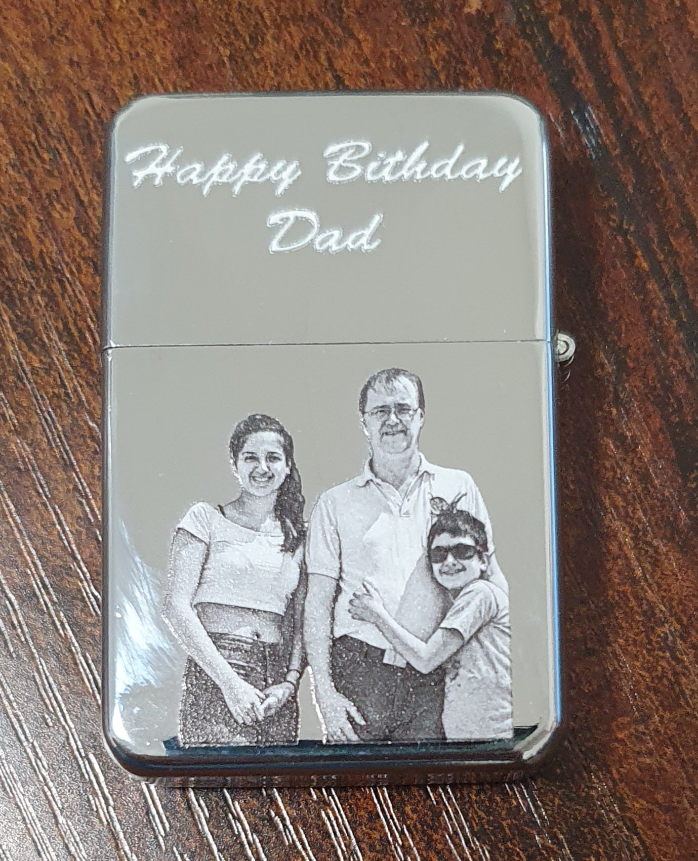 A custom engraved metal lighter with a brushed silver finish, featuring the text 'Happy Birthday Dad' and a black-and-white family photo of three on the front, resting on a dark wood surface.