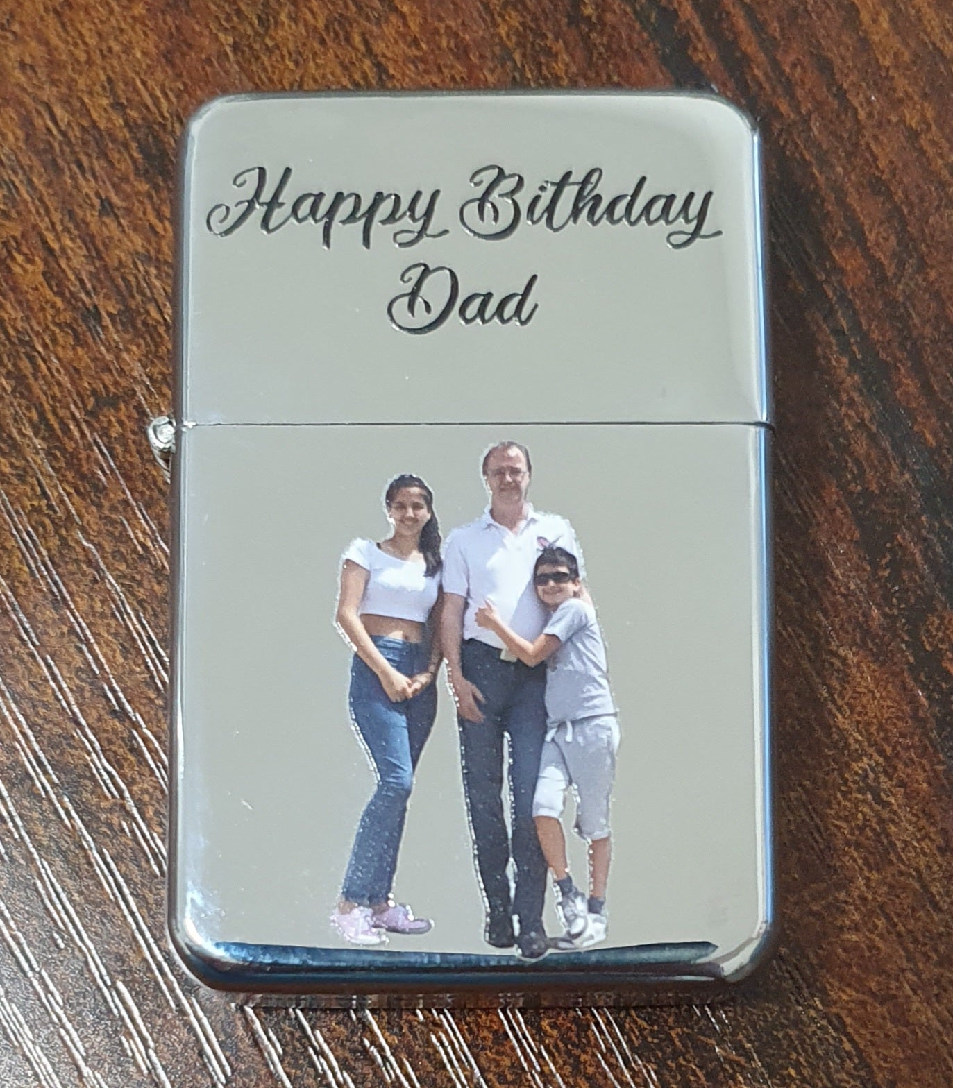 A custom-engraved chrome lighter resting on a dark wood table. The lighter features the text 'Happy Birthday Dad' in black cursive script on a brushed silver background, with a custom photo of a father and two children printed below.