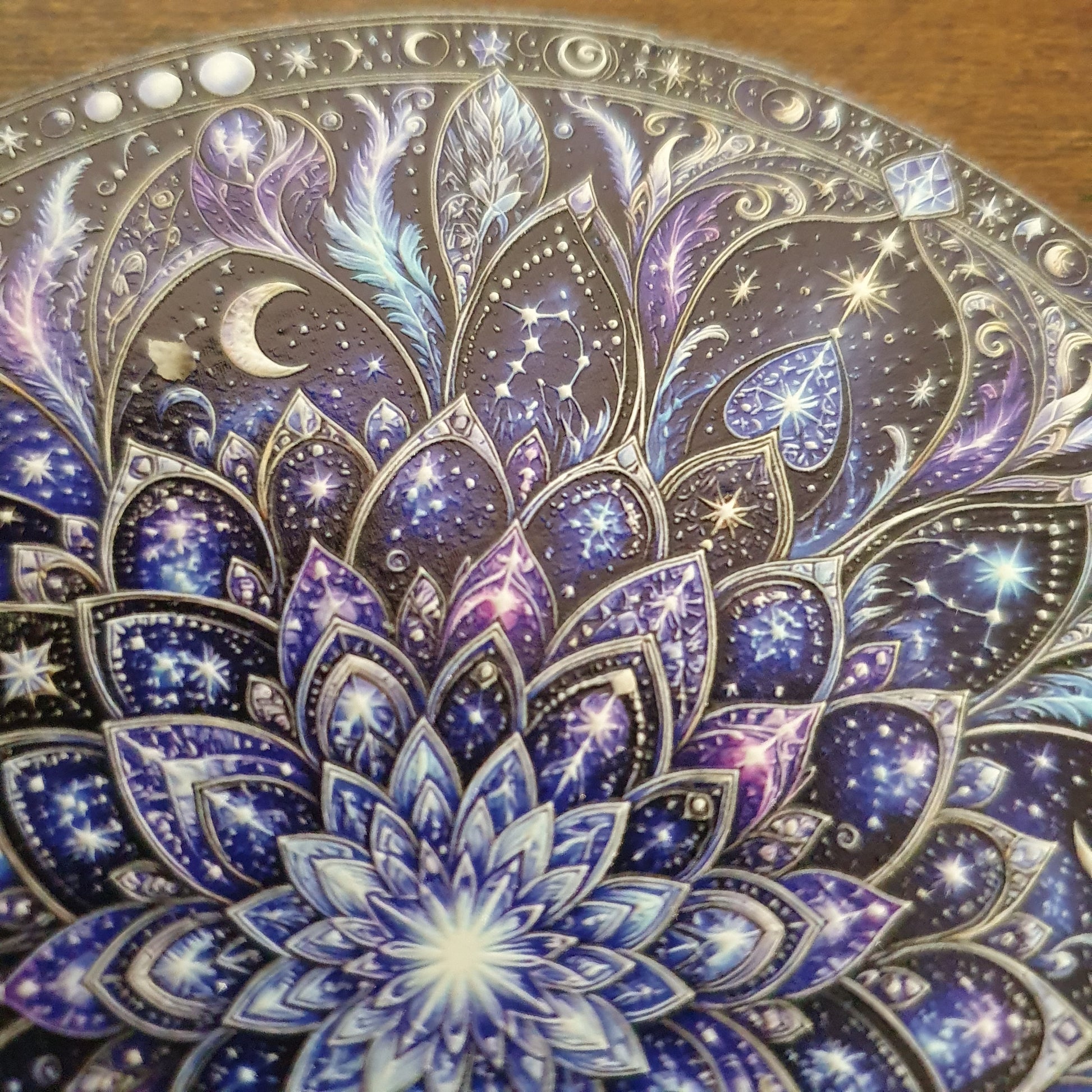 A close-up of an ornate circular decorative plate or tray featuring a detailed celestial mandala design in deep blue, purple, and silver tones, with stars, crescent moon, and feather motifs.
