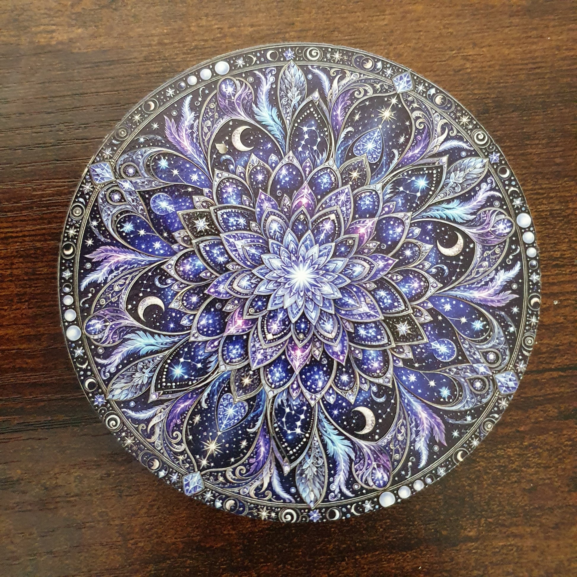 A round decorative item, likely a compact or box lid, featuring an intricate hand-painted mandala design in shades of purple, blue, black, and white, set against a dark brown wooden background.