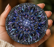 A hand holds a round decorative item featuring an intricate celestial mandala design in deep blues, purples, and black with white star-like speckles.