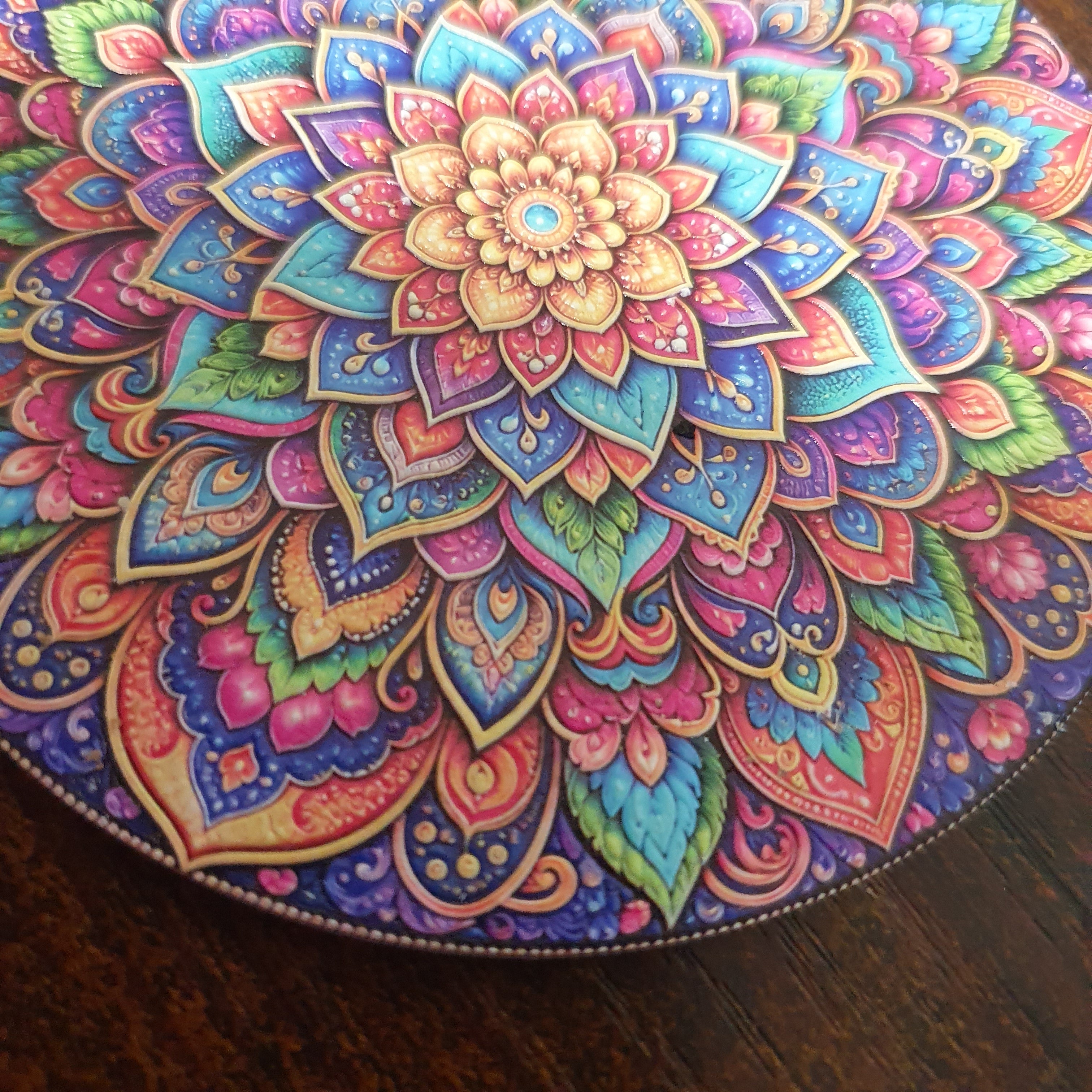 A round decorative tray or plate featuring a highly detailed, colorful mandala design in shades of blue, pink, purple, green, and gold, photographed on a dark wooden surface.
