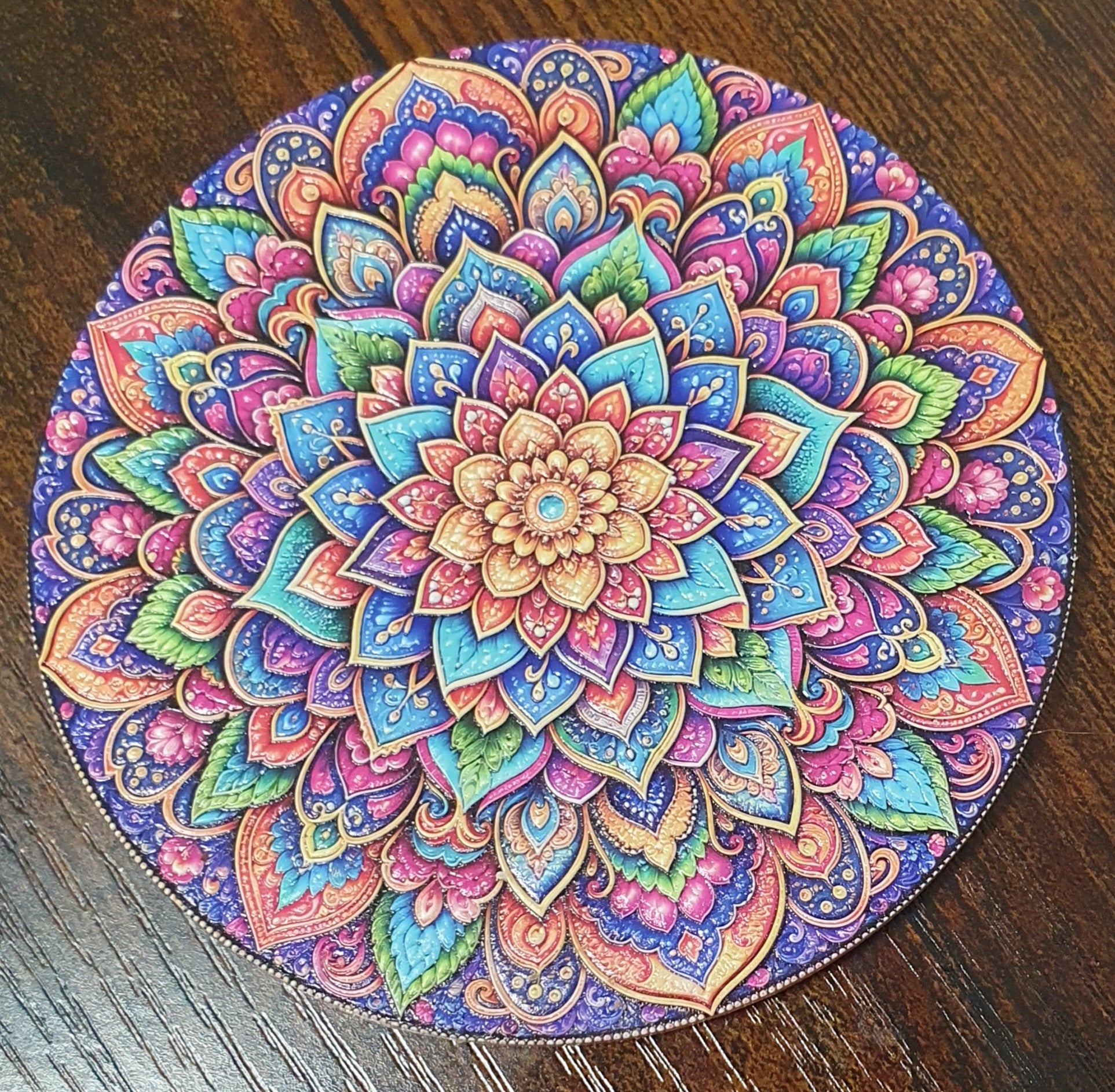 A round coaster featuring a highly detailed, colorful mandala design, placed on a dark brown wooden surface.