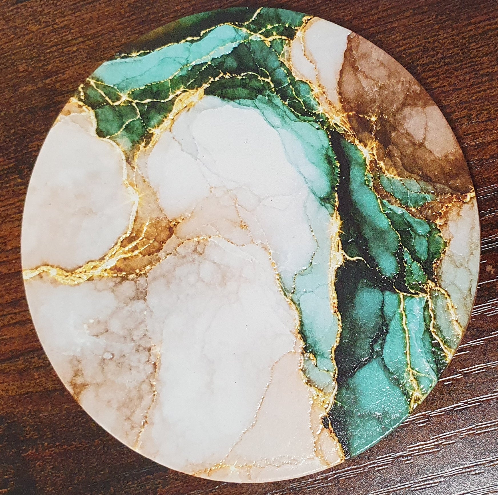 A round marble-patterned coaster with green, white, and brown veining and gold metallic accents, shown on a dark wood surface.