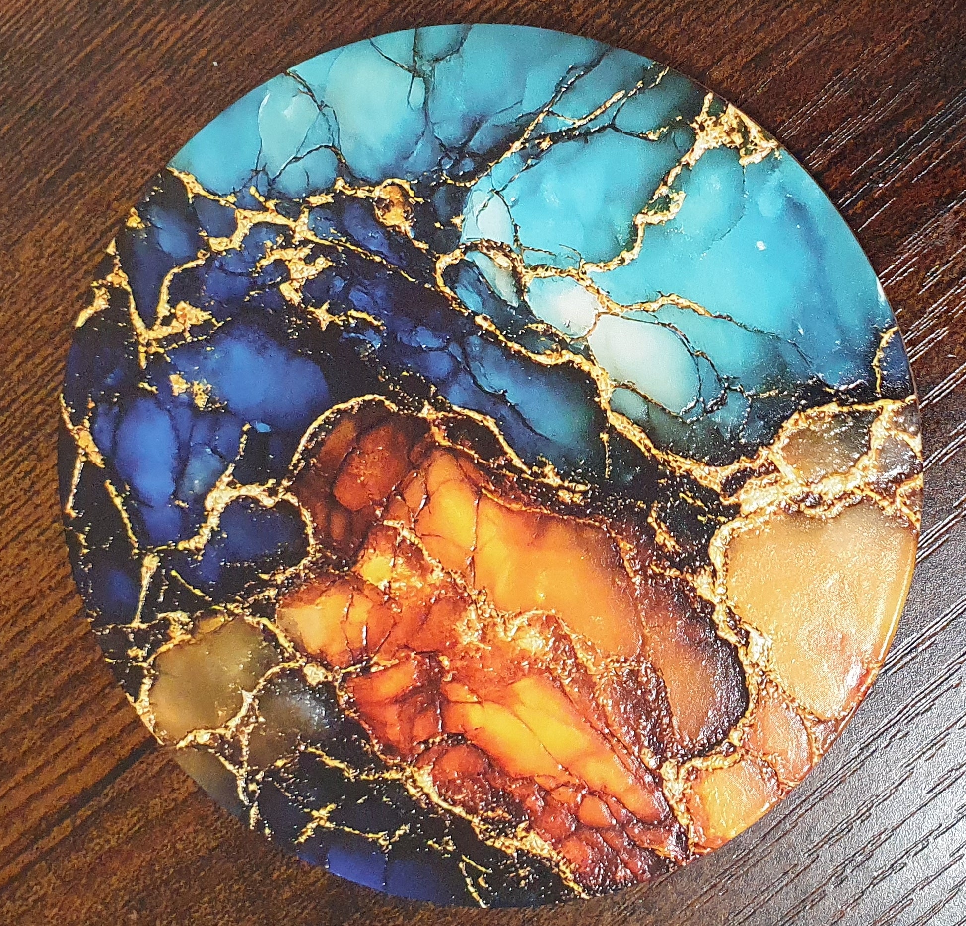 A round coaster featuring a vibrant marbled pattern with turquoise, deep blue, orange, and gold veins, displayed on a dark wood surface.