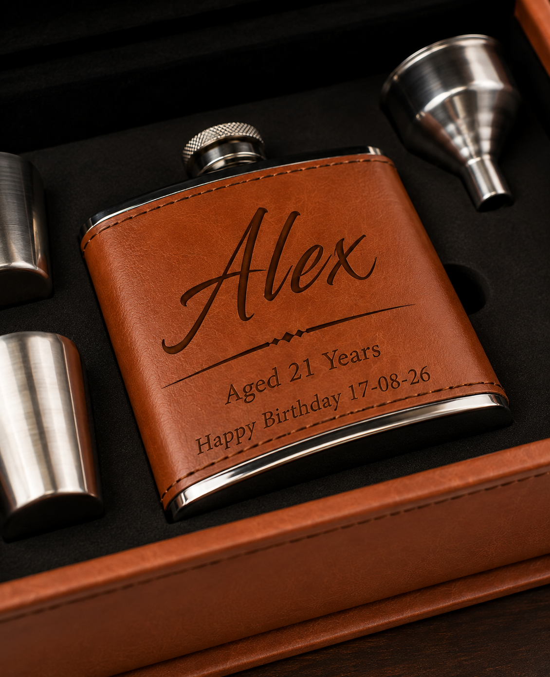A personalized brown leather flask with silver metal trim and a screw-top cap, shown inside a gift box with matching silver accessories. The flask is engraved with the name 'Alex', 'Aged 21 Years', and a birthday date.