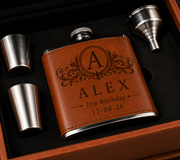 Personalized brown leather flask with engraved monogram and birthday text, displayed in a gift box with two metal flasks and a funnel.