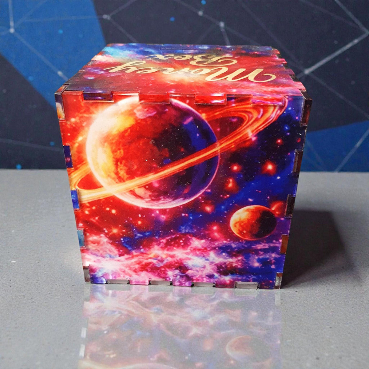 Close-up of galaxy money box featuring a large ringed planet and bright cosmic background in vivid red, orange and blue colours. Modern acrylic coin bank with slot on top for saving coins.